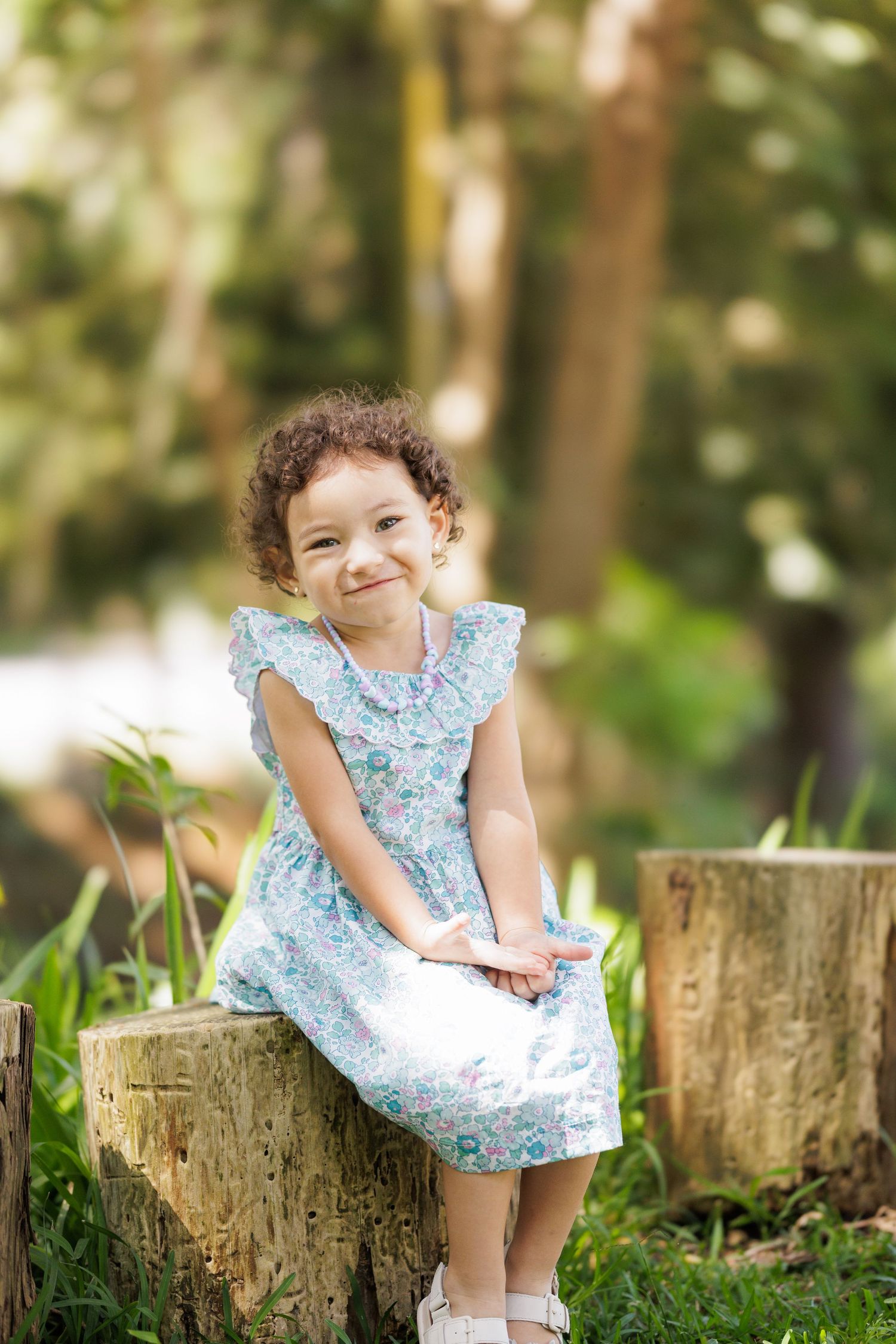 A child in a blue floral dress sits on a tree stump in a sunny garden setting.