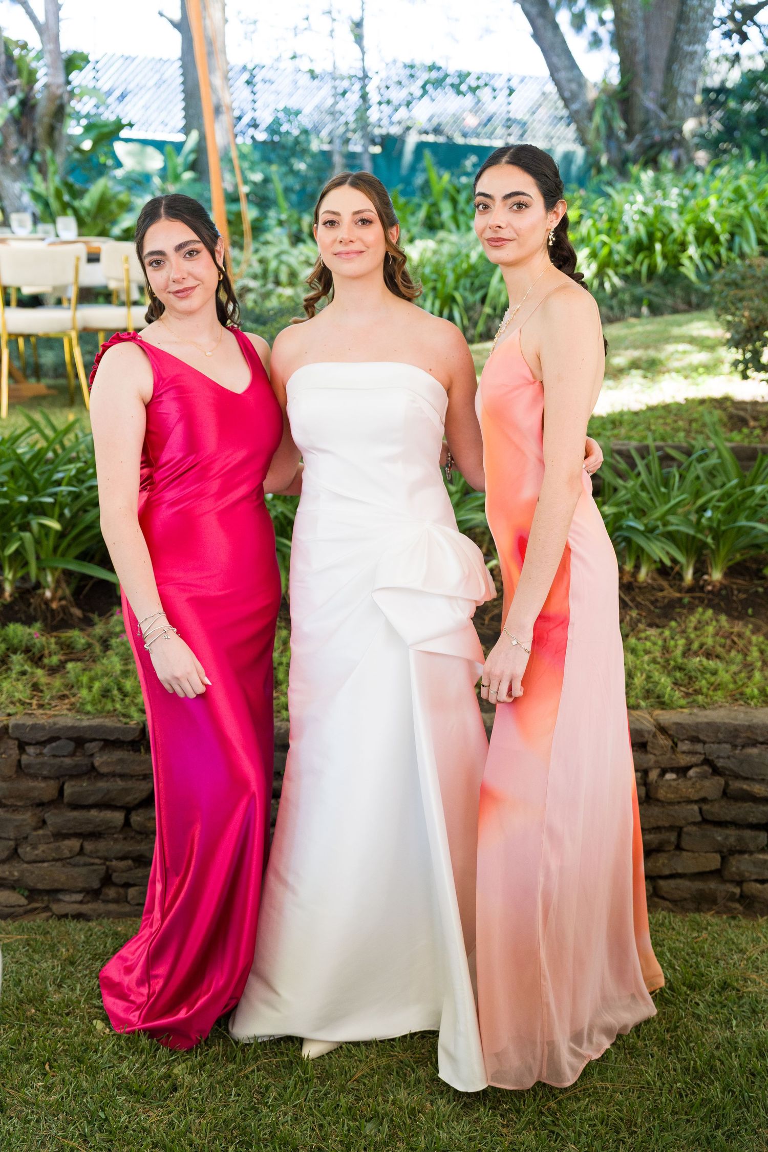 Bridal party in red and coral dresses pose on grass near stone wall.