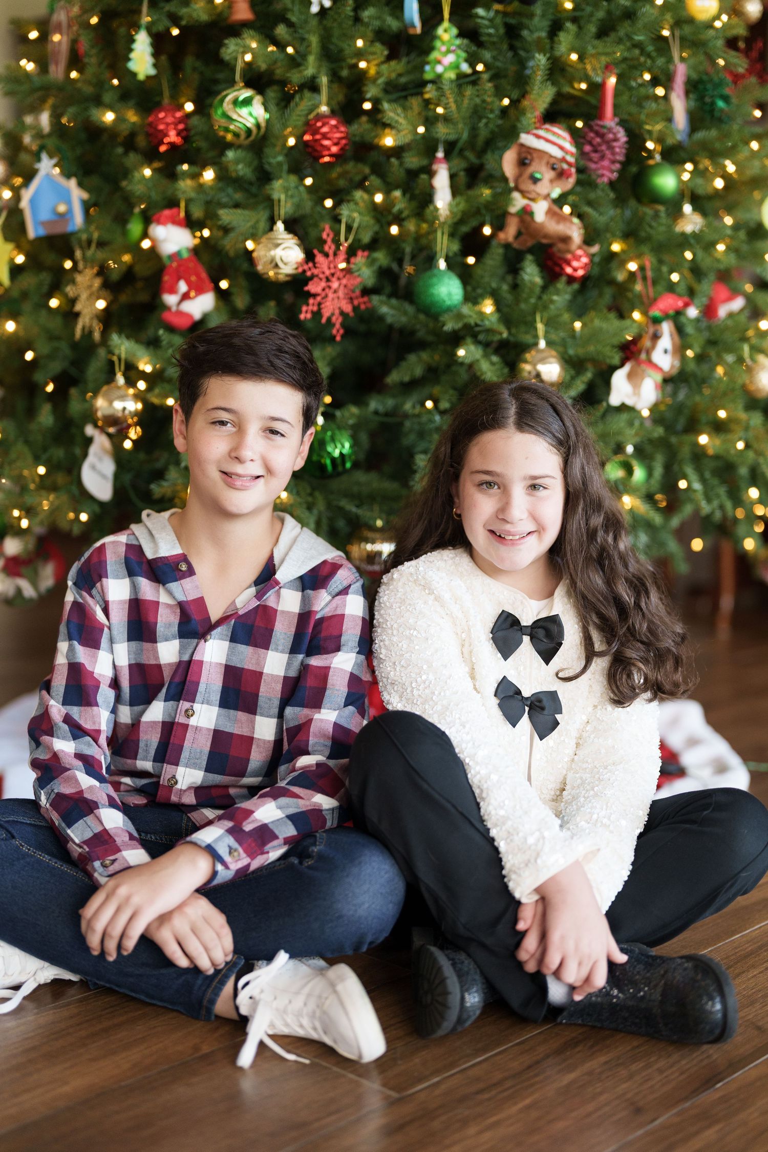 Siblings pose together on hardwood floor with decorated Christmas tree in background.