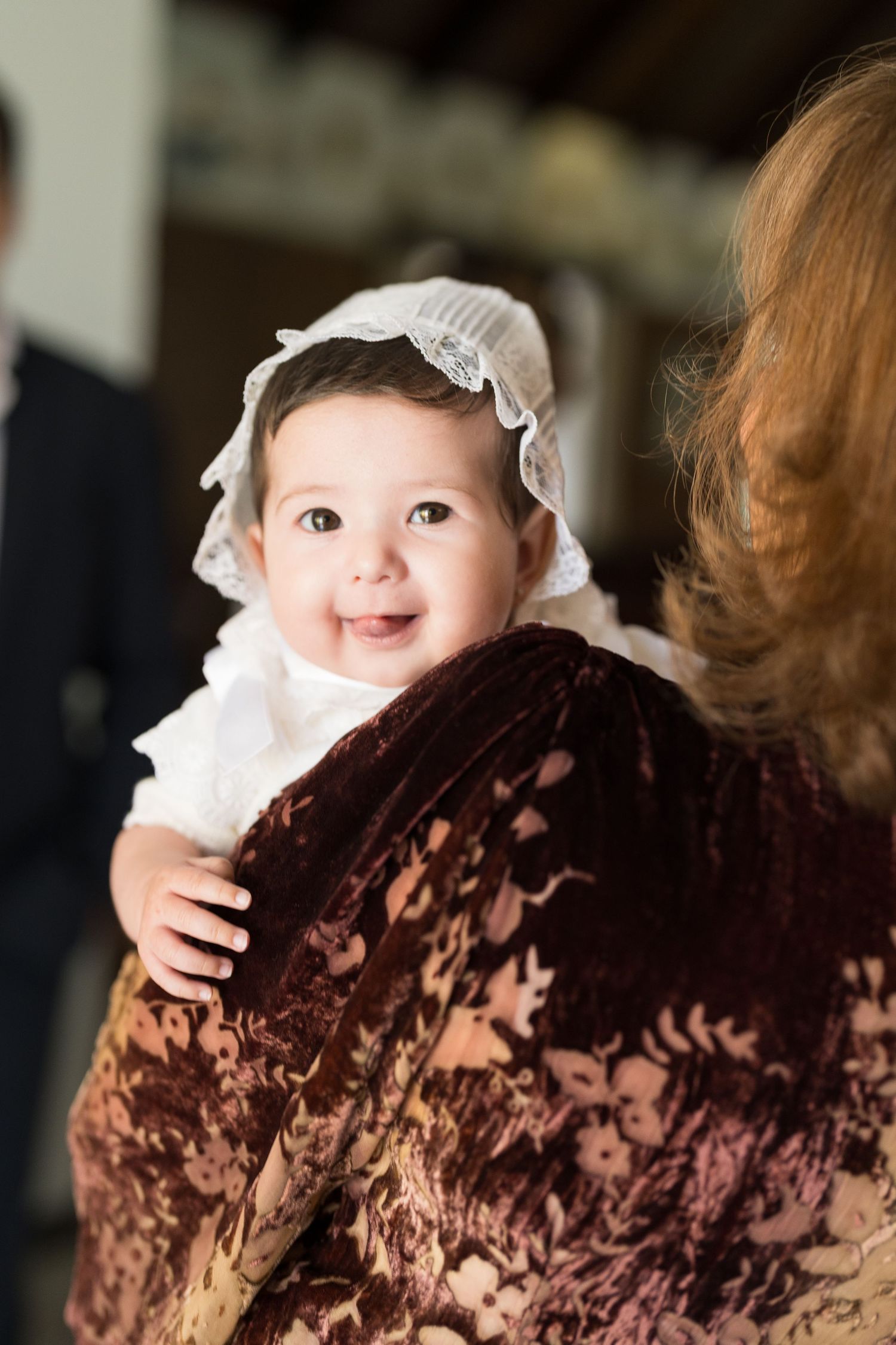 Baby in white baptismal gown peeking over someone's shoulder wearing burgundy velvet.