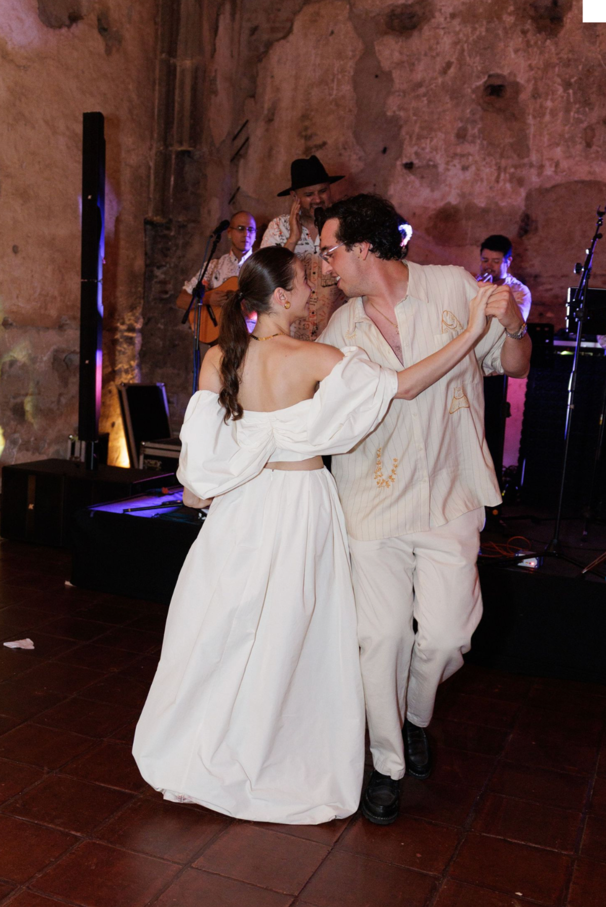 A couple dances together in white outfits under colorful lighting in a rustic venue.