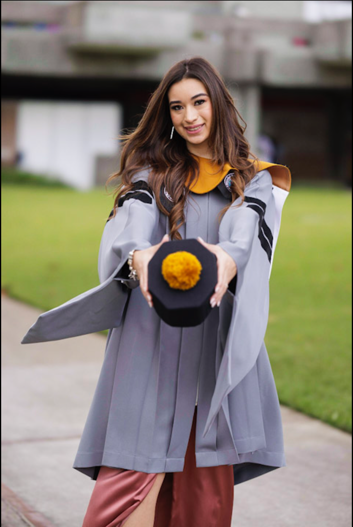 Graduate in grey academic regalia and yellow stole holds up graduation cap while smiling.