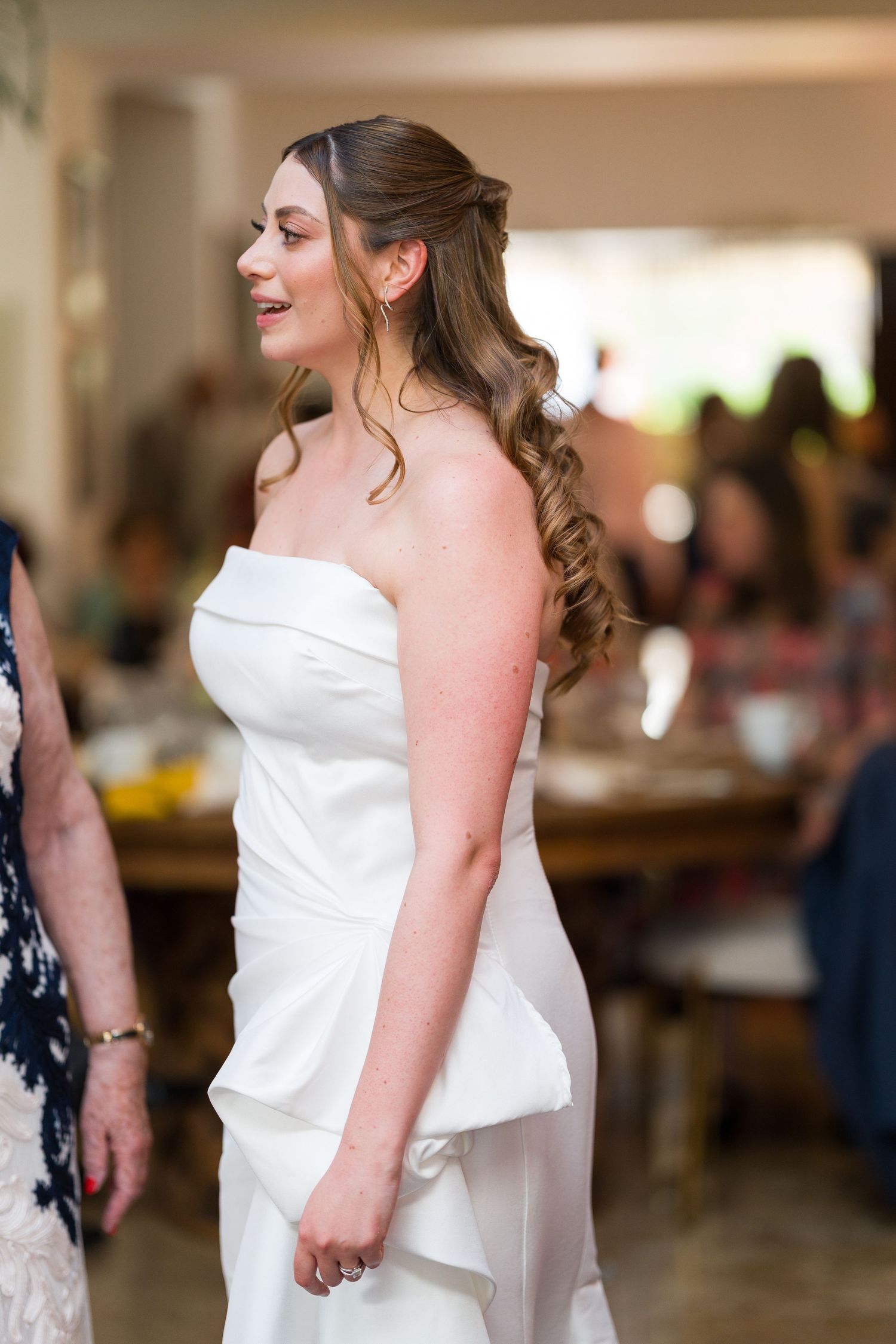 A bride in an elegant white strapless wedding gown stands in an indoor venue with brick walls.