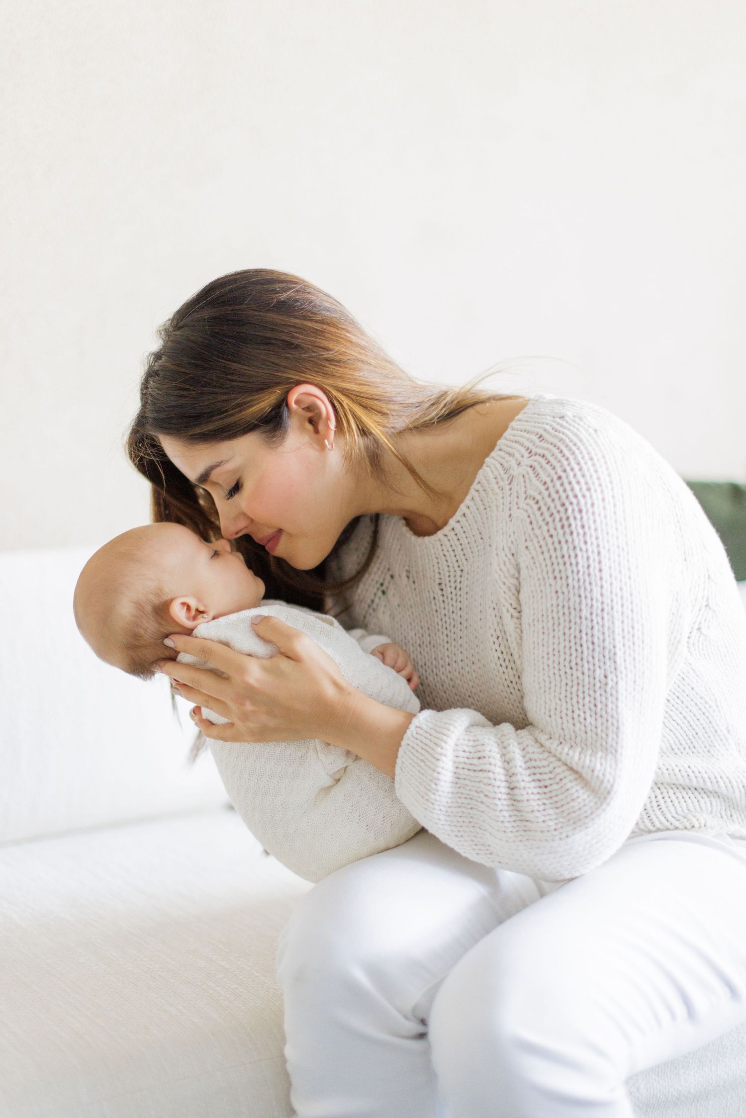 Mother in white sweater cuddling with baby on white bedding.