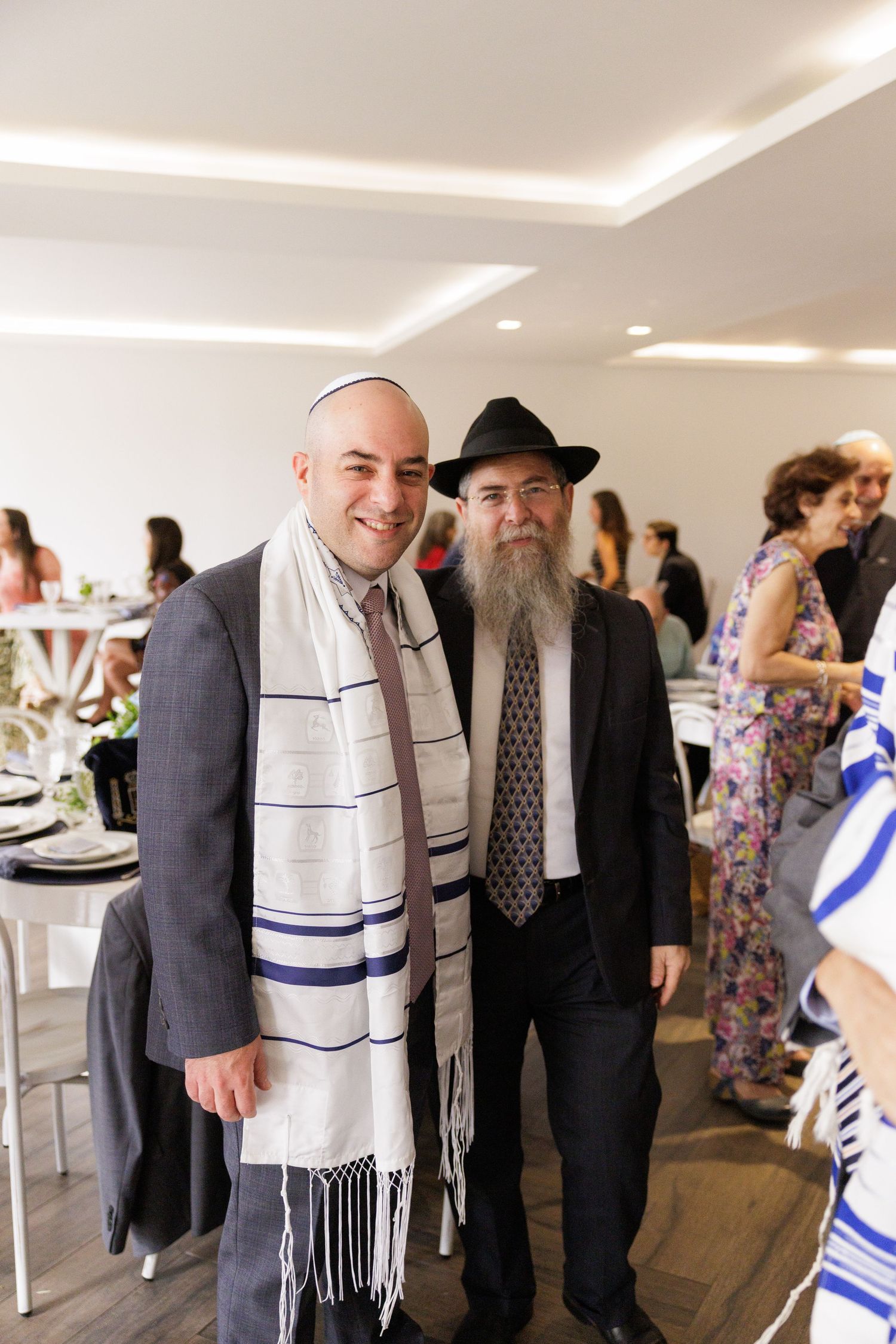 Two men wearing formal attire and prayer shawls stand together at an indoor celebration.