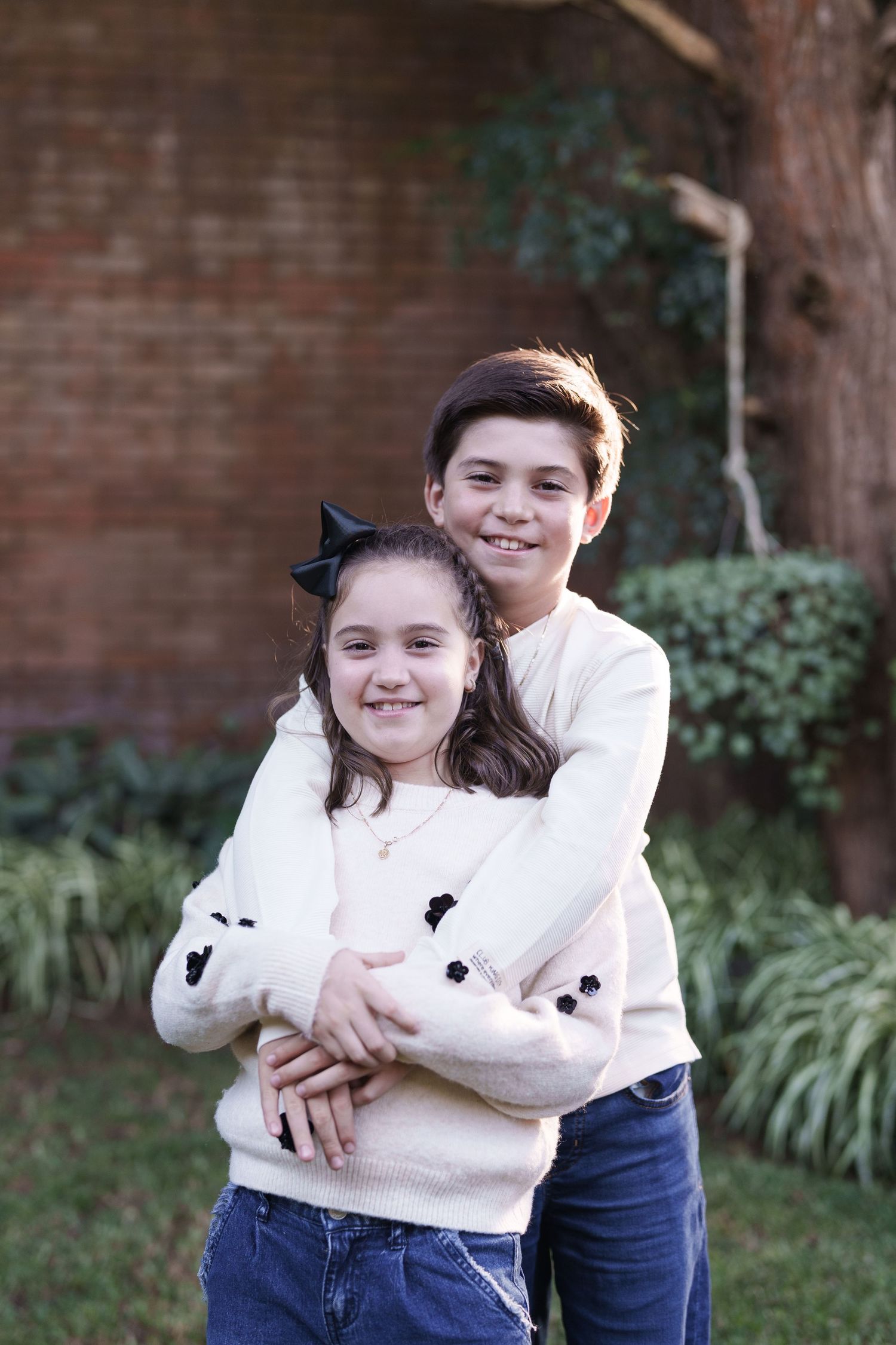 Two siblings sharing a warm embrace against a brick wall backdrop with greenery.