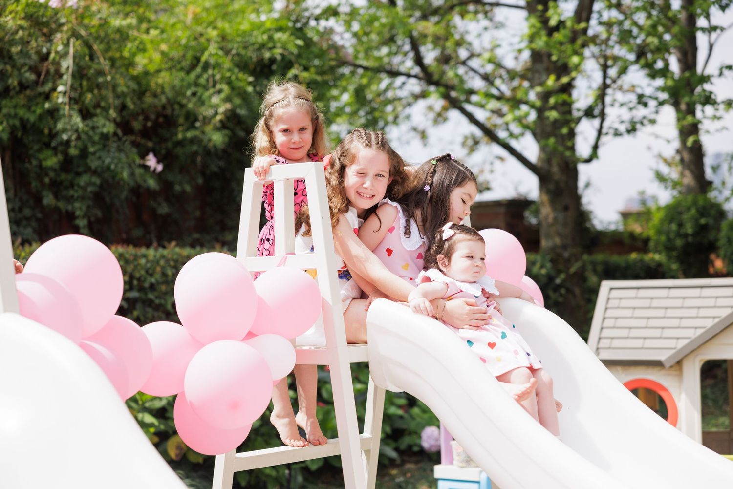 Pink balloons decorate a white slide in an outdoor garden setting during a festive celebration.