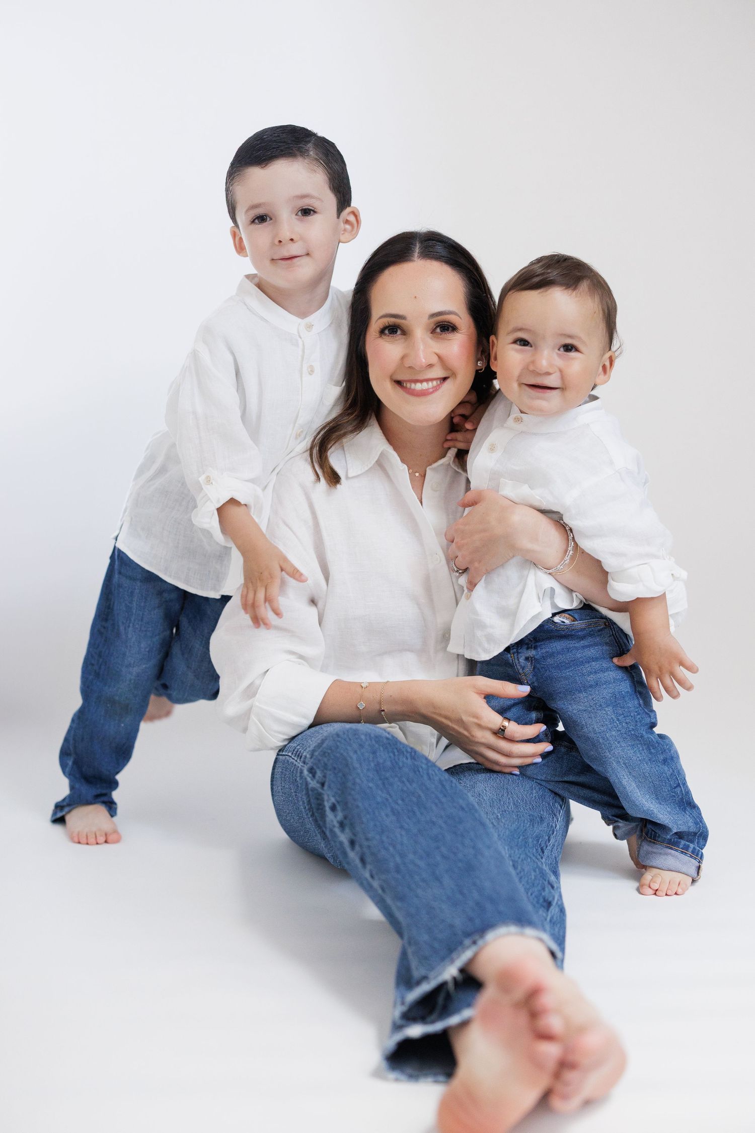 A smiling mother in white shirt and jeans sits on floor with two young boys in matching outfits on white background.