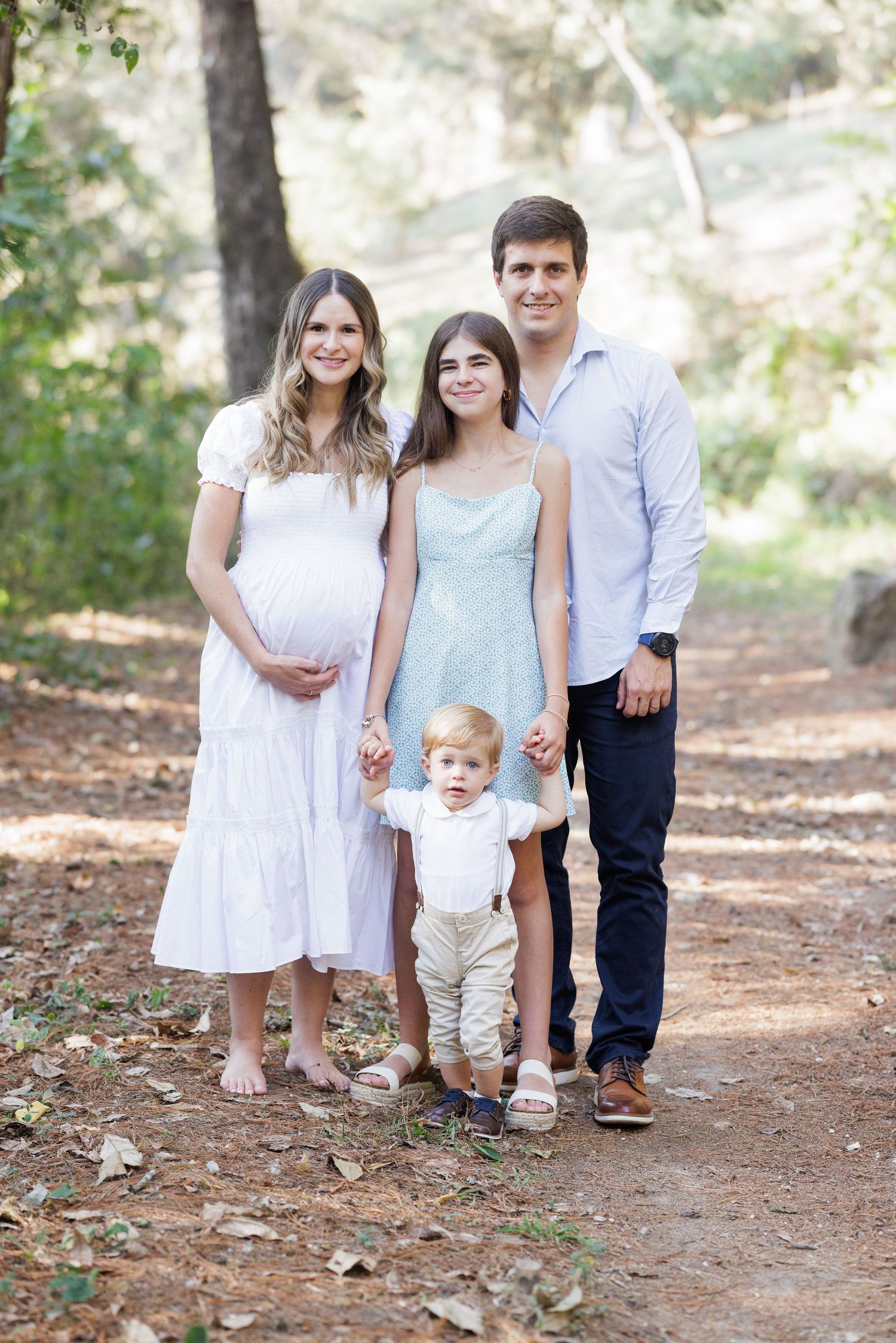 A family group walks together on a wooded path wearing coordinated light-colored outfits.