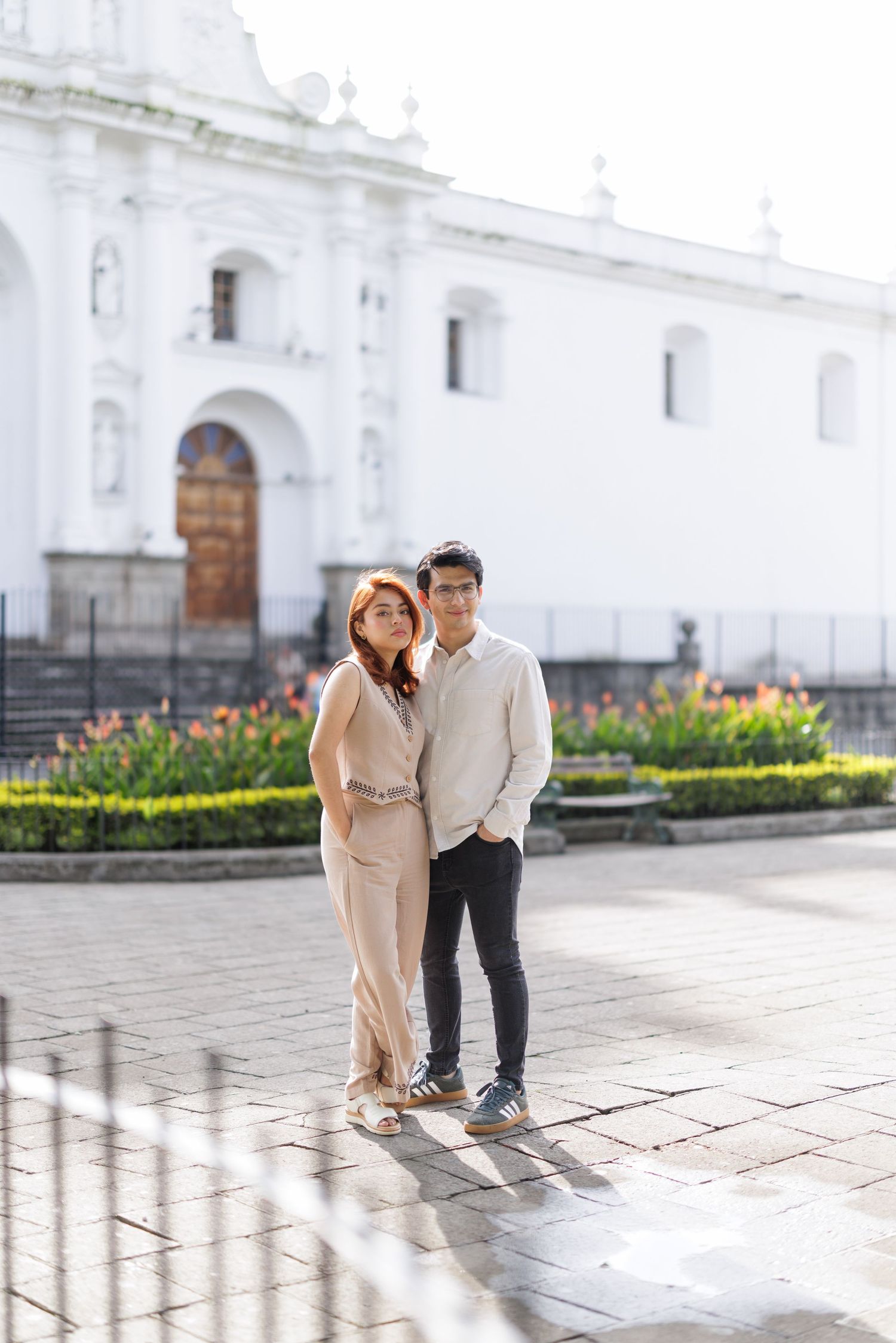 Couple poses playfully on stone plaza in front of white colonial church.