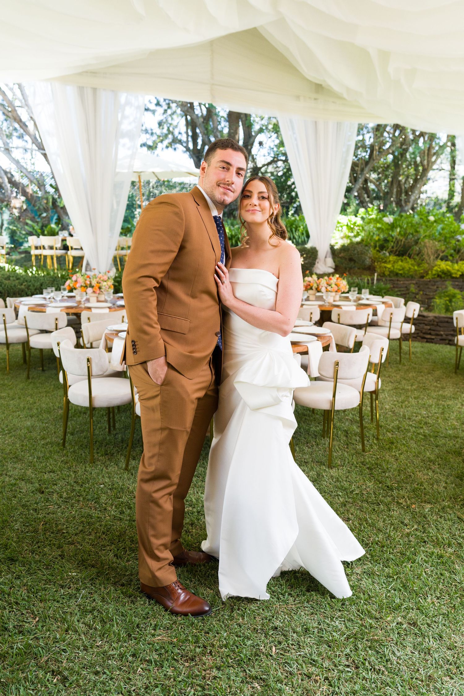 Wedding couple poses playfully on grass with white reception tables in background.