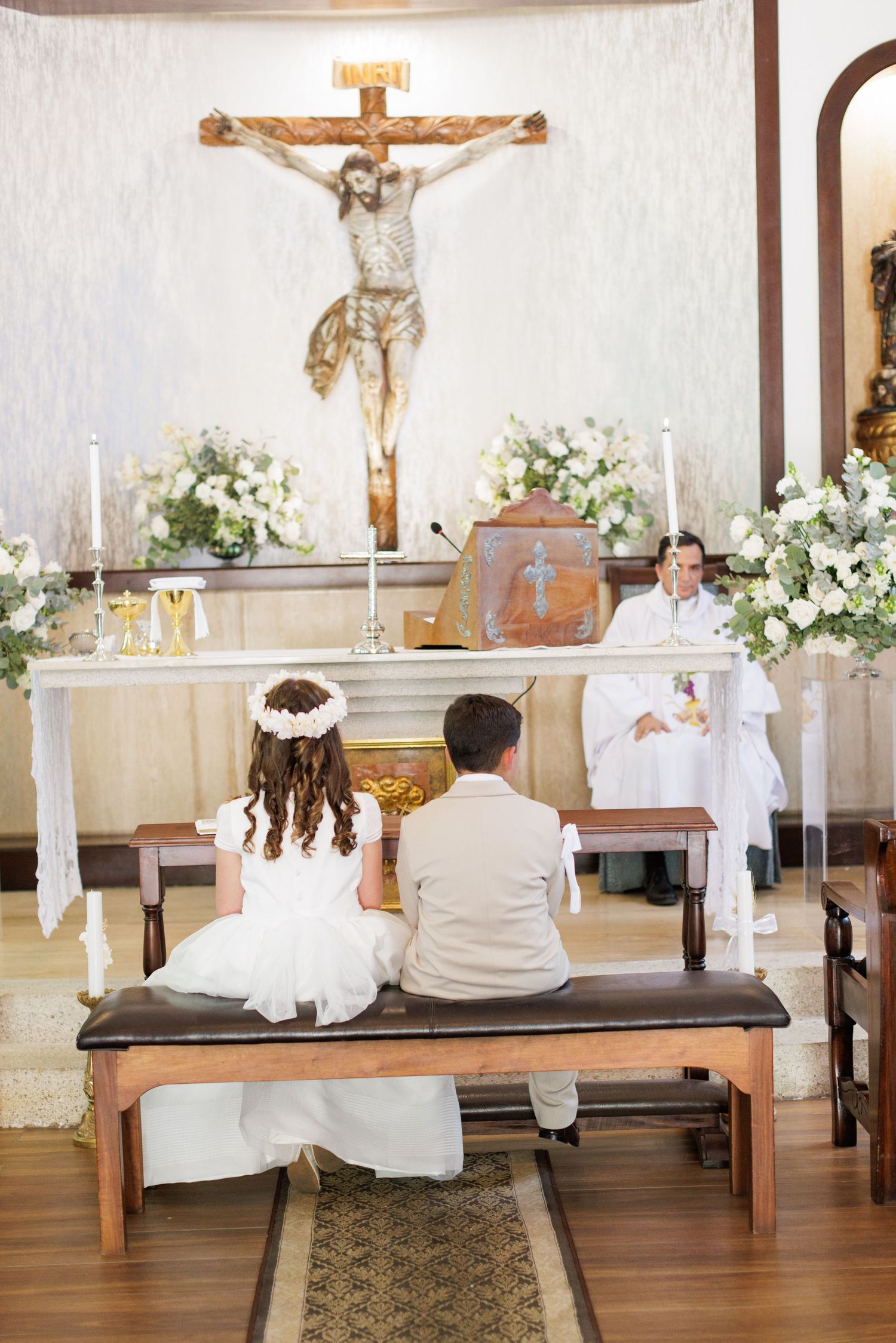 Interior of Catholic church with crucifix, altar, and white floral arrangements during ceremony.