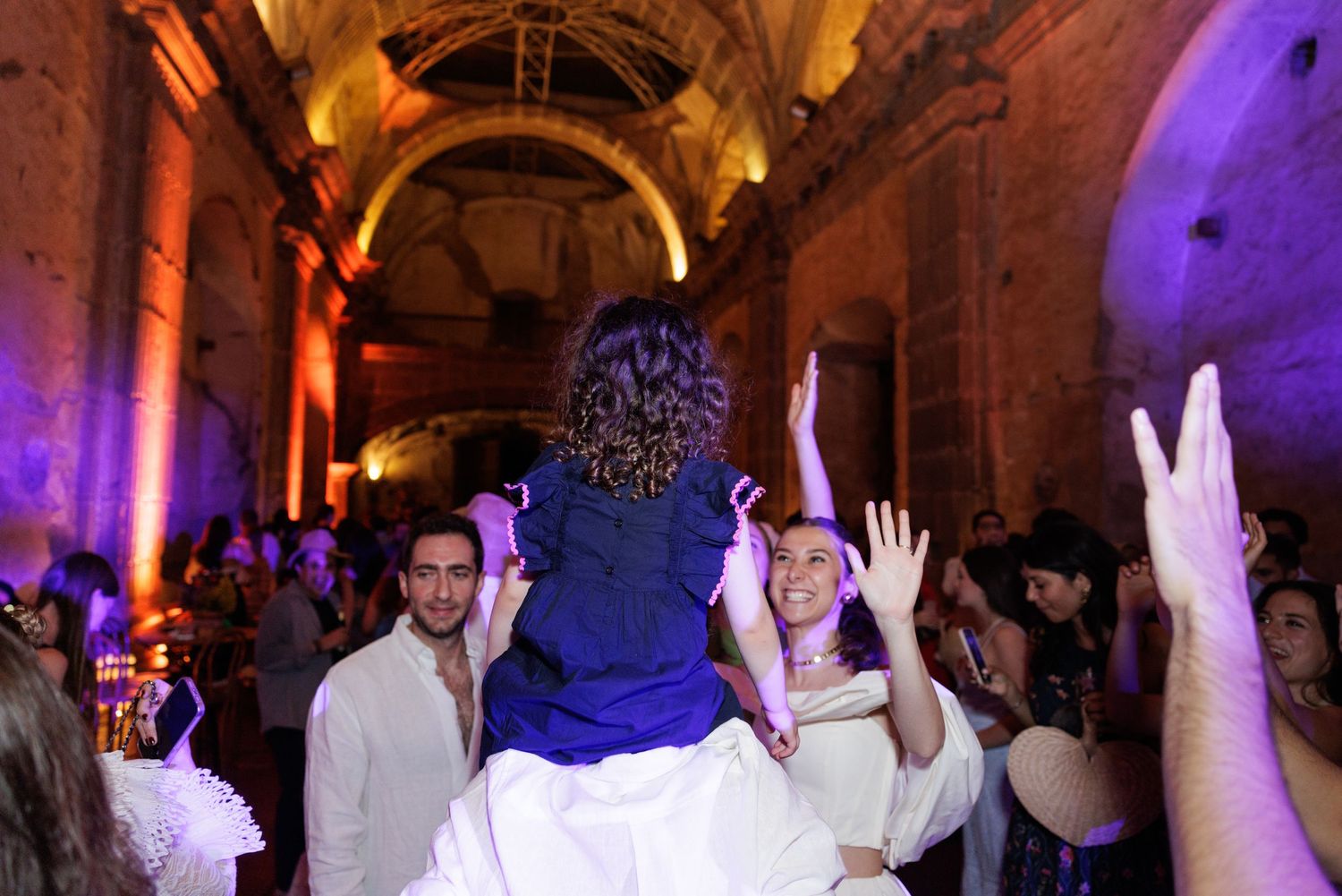 Celebratory dance scene with raised hands under dramatic orange and purple lighting in historic venue.
