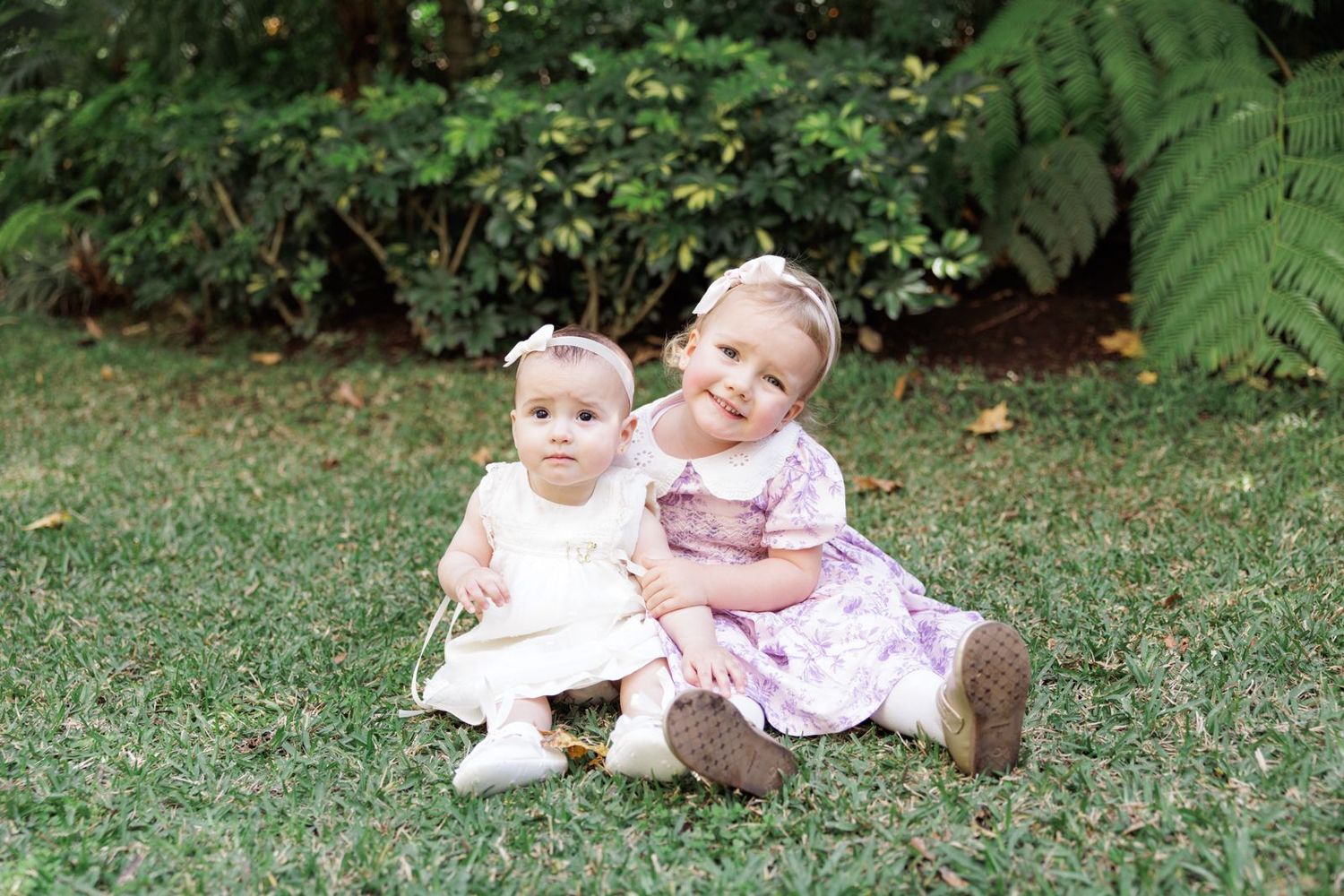 Two young children in light colored dresses sitting together on grass with ferns in background.