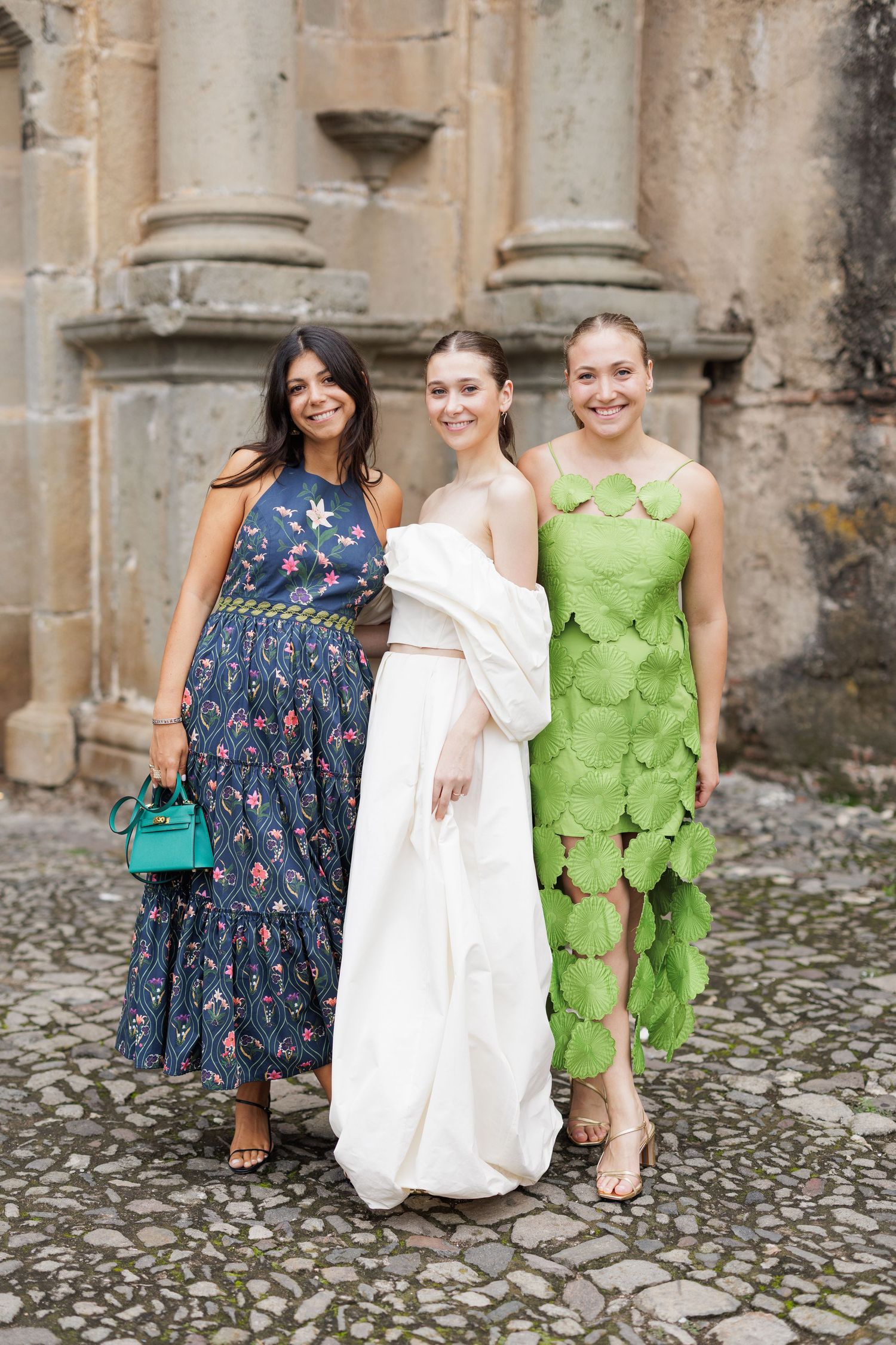 Three friends in colorful dresses posing together on cobblestone ground near stone architecture.