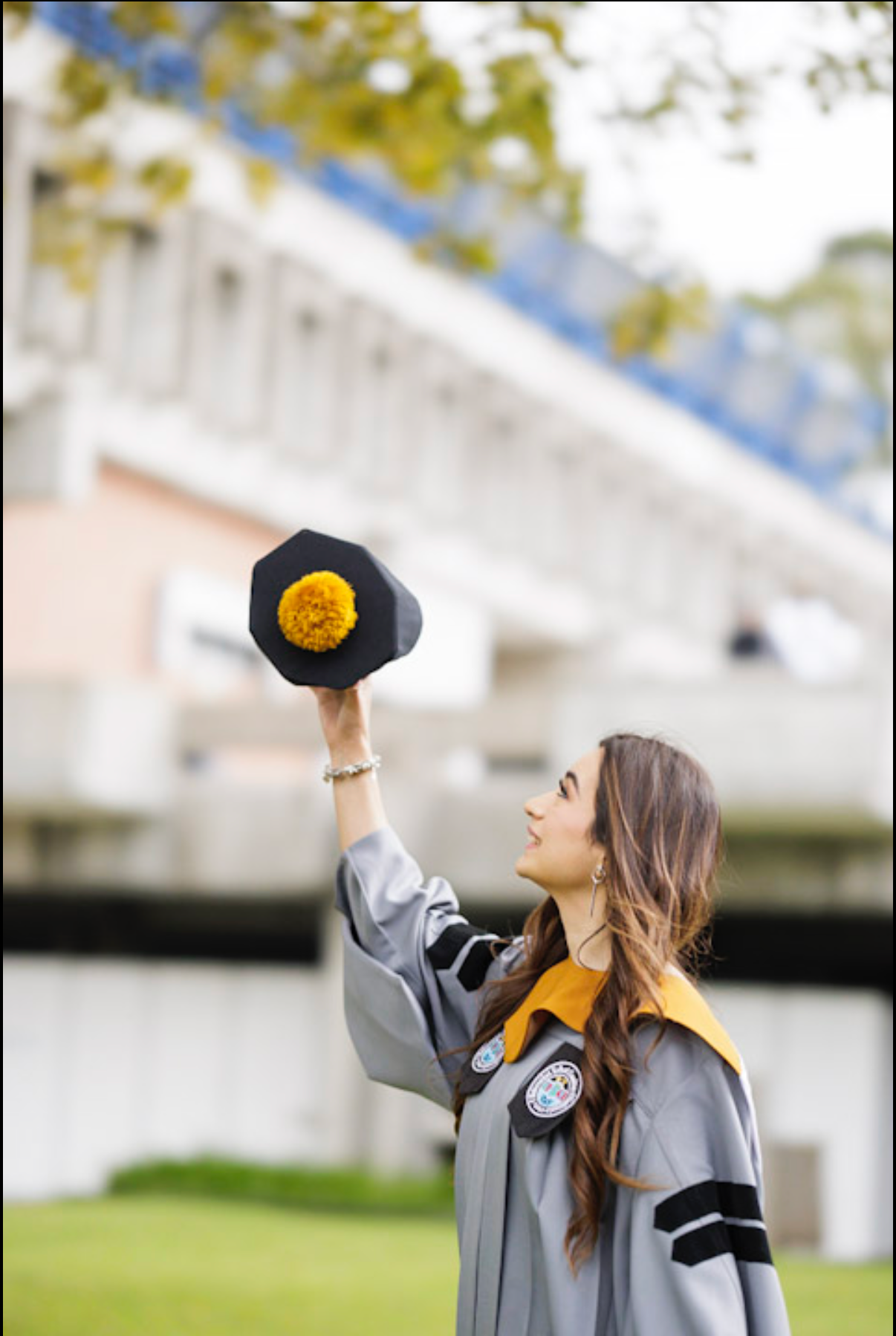 Graduate in grey academic regalia raises cap triumphantly against blurred building background.