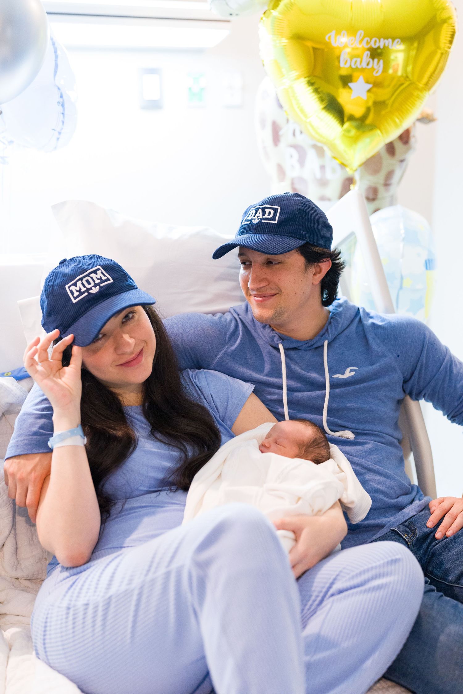 Couple wearing matching blue outfits and baseball caps pose with newborn under yellow celebration balloons.