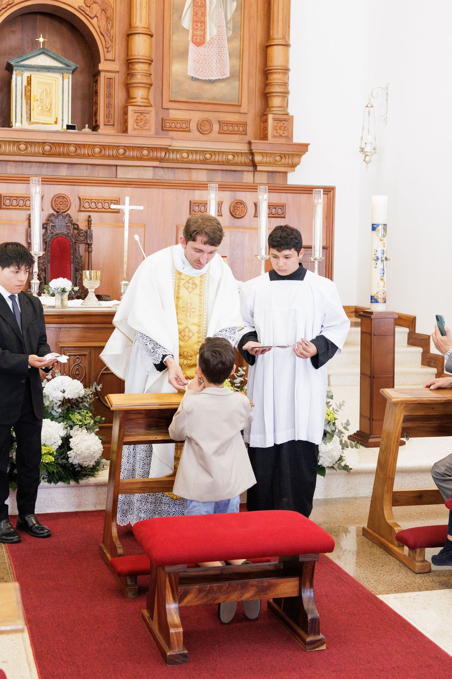 Church altar servers and clergy during traditional Catholic ceremony with red carpet.