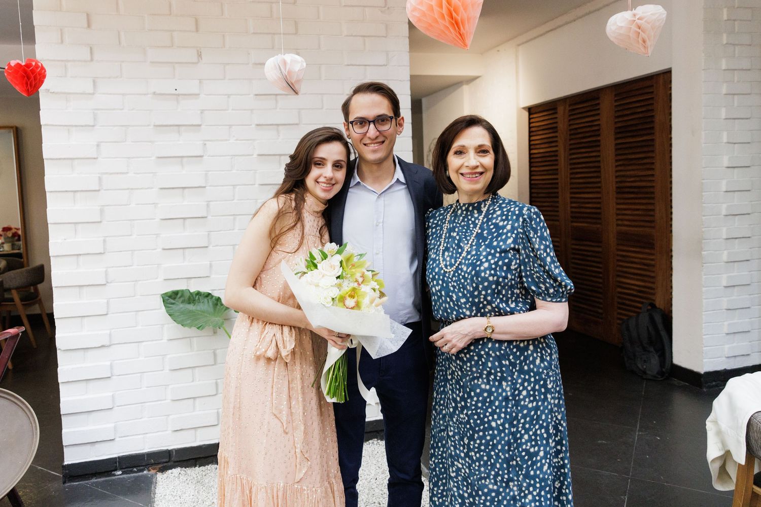 Wedding guests in formal attire pose together against a white brick wall.