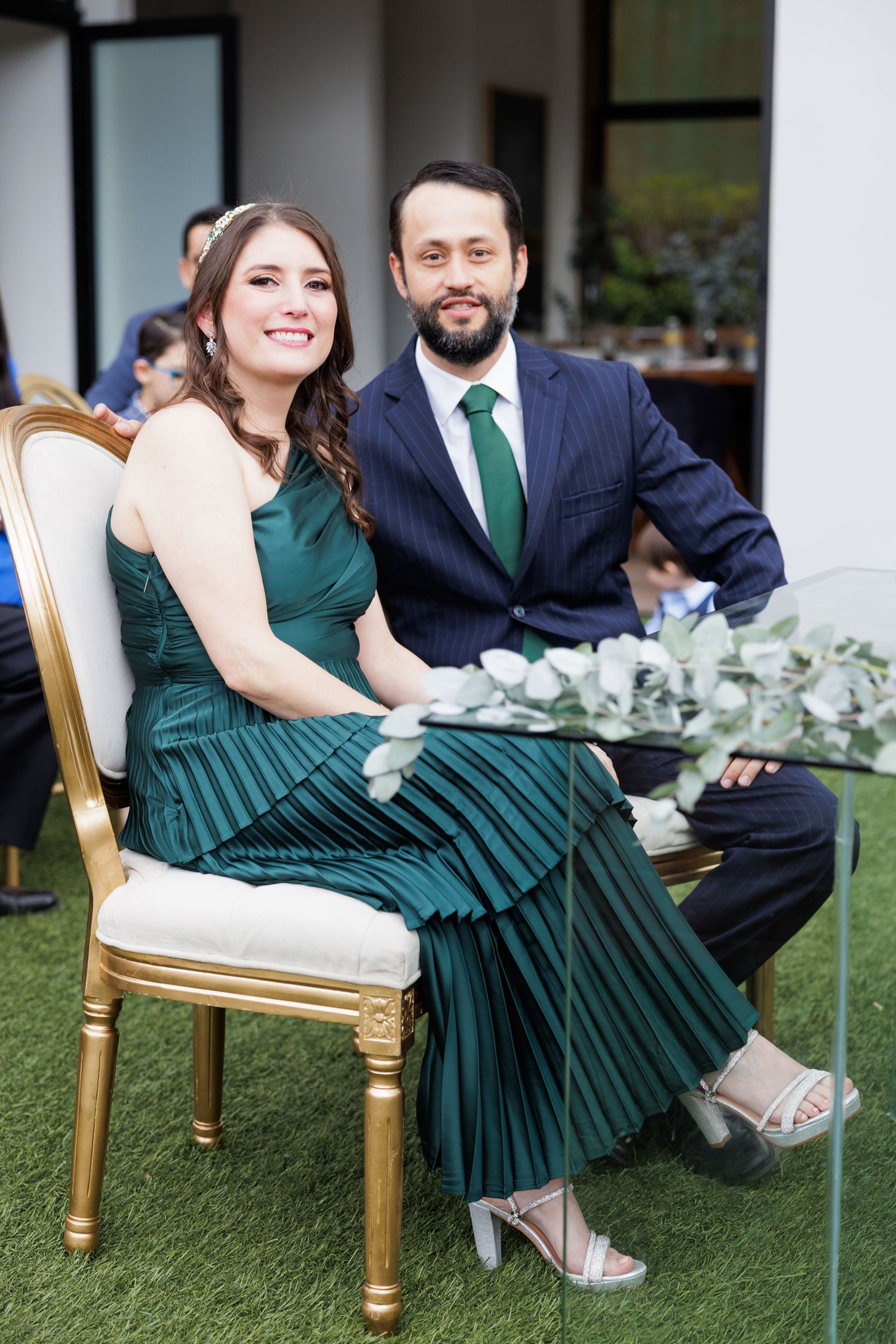 Couple relaxes on gold chairs during outdoor event with emerald green dress and navy suit.