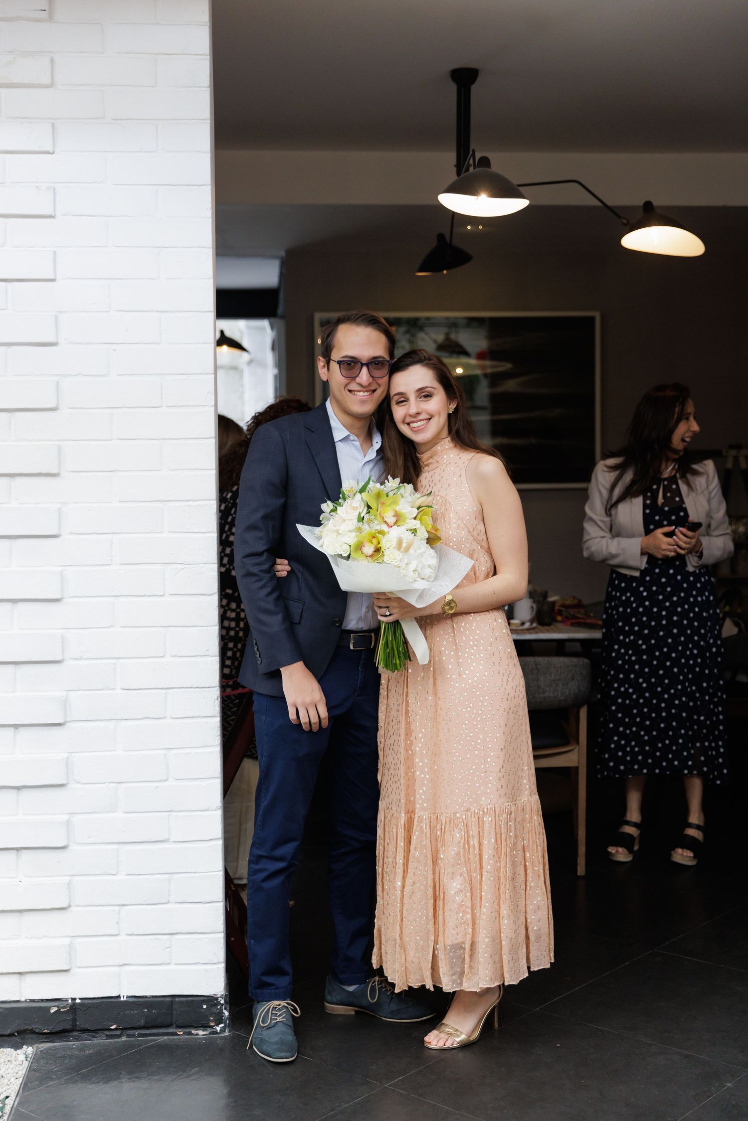 A couple poses with a bouquet against a white brick wall at an indoor venue.
