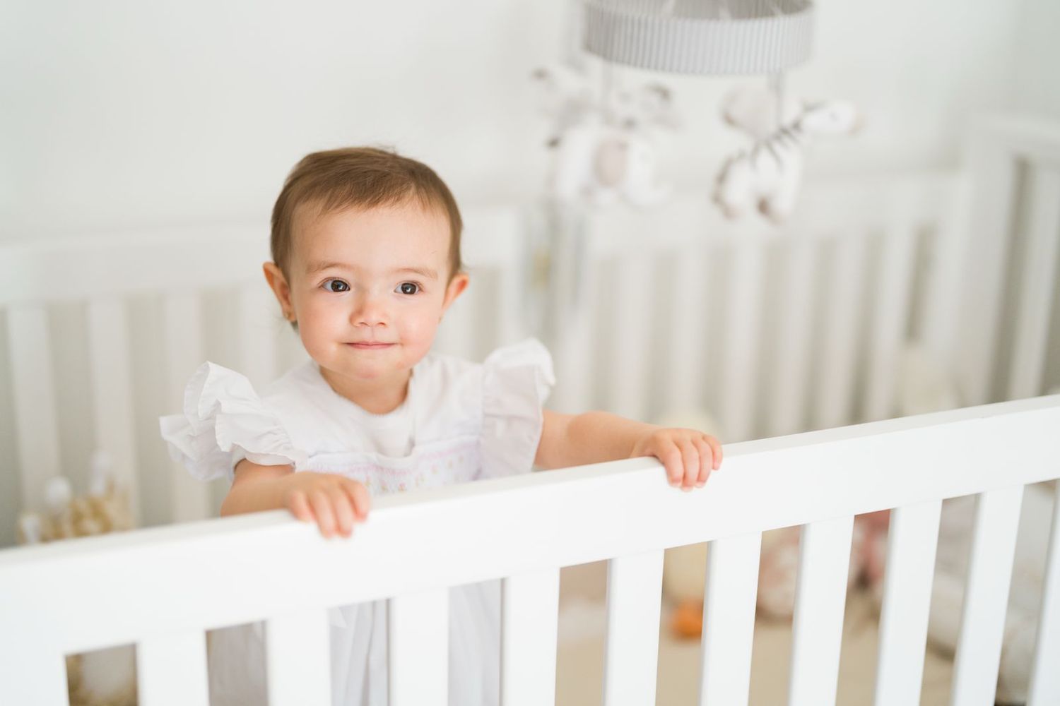 Toddler in white outfit standing in white crib looking at camera.