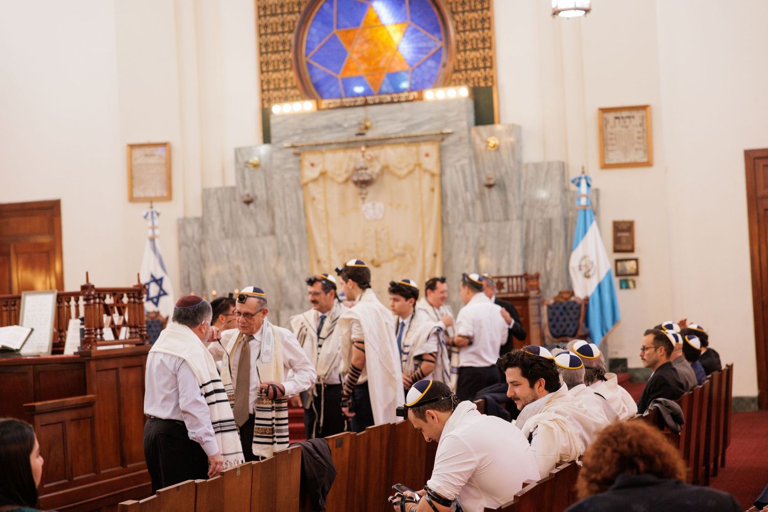 Interior of a synagogue showing wooden pews and stained glass windows during a prayer service.