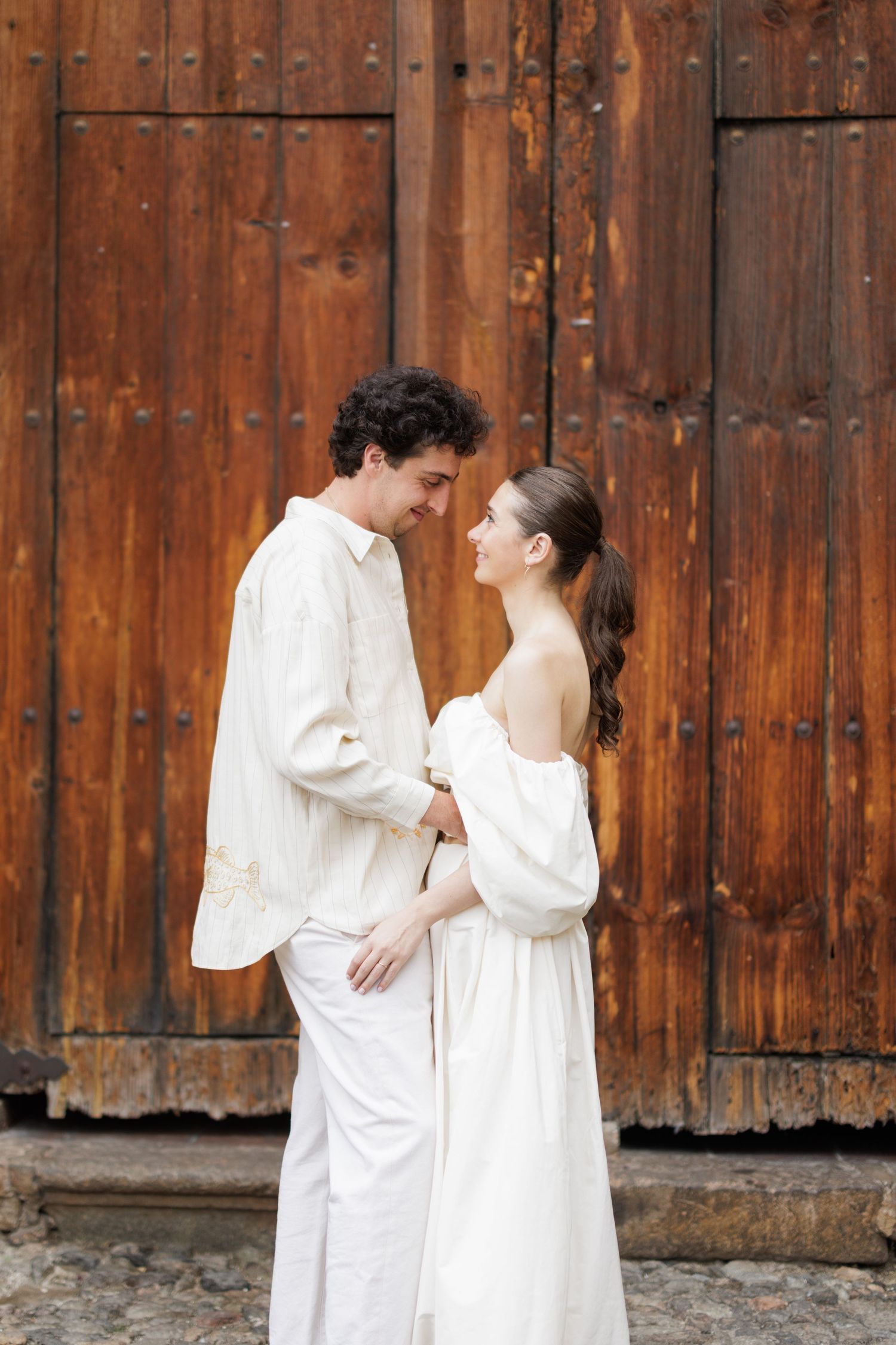Couple in flowing white outfits against rustic wooden wall background.