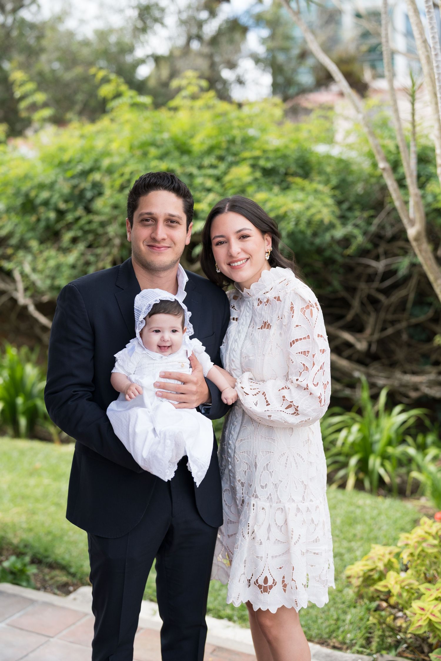 Family gathered outdoors for baptism photos in formal attire with lush garden background.