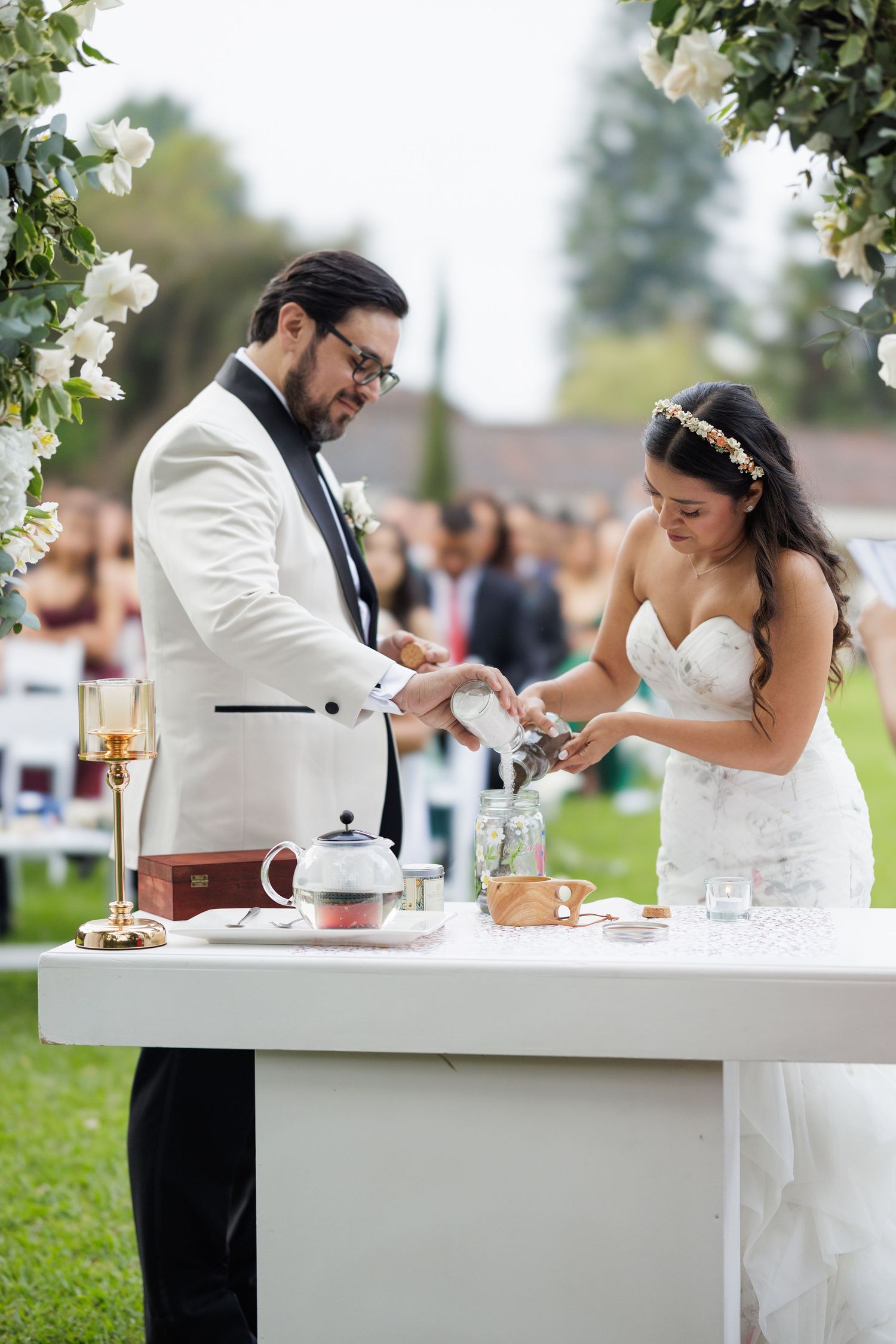 Newlyweds enjoy a tea ceremony at an elegant white-decorated outdoor reception table.