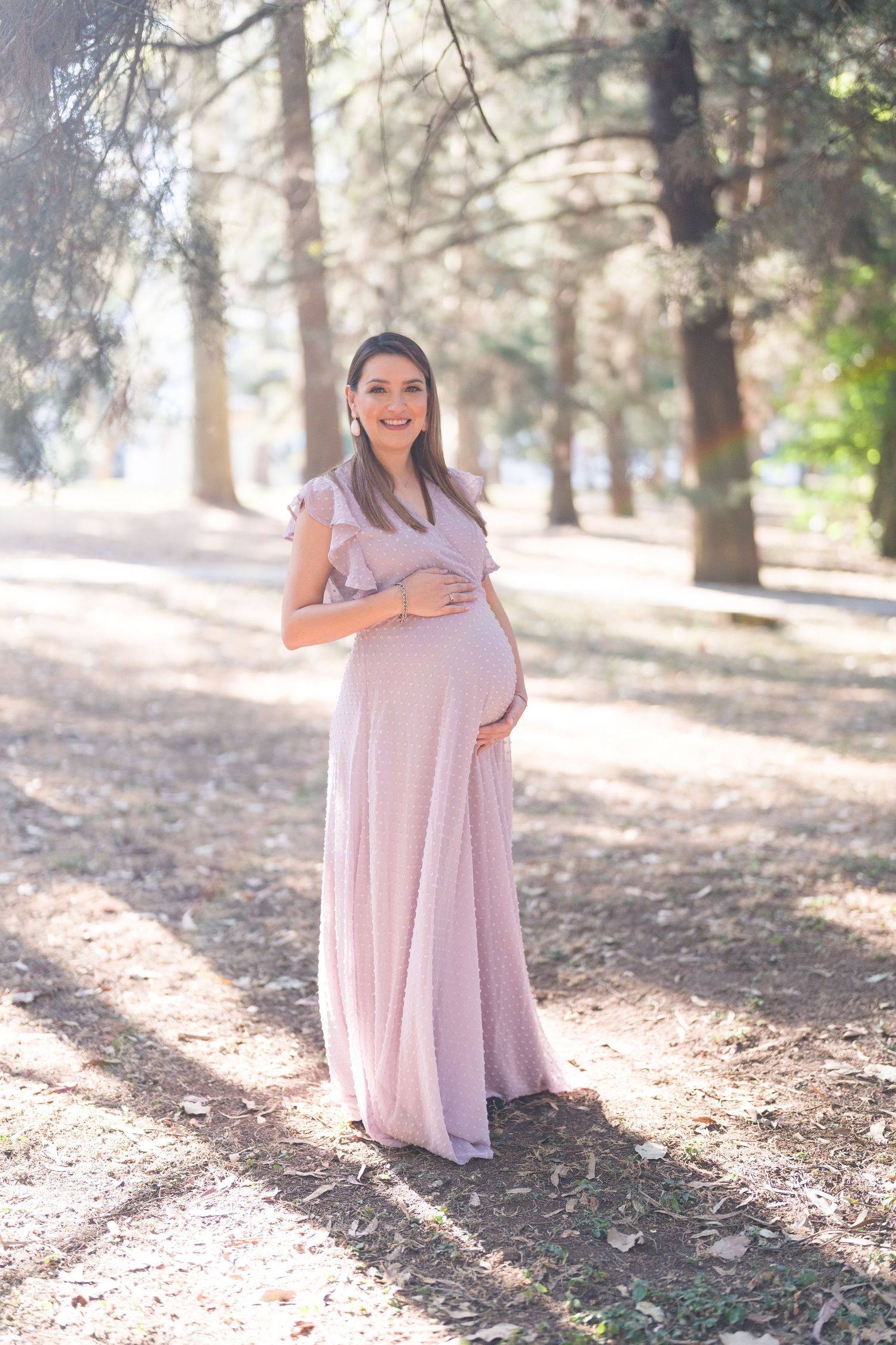 Expectant mother in a flowing mauve dress poses gracefully in a wooded setting.