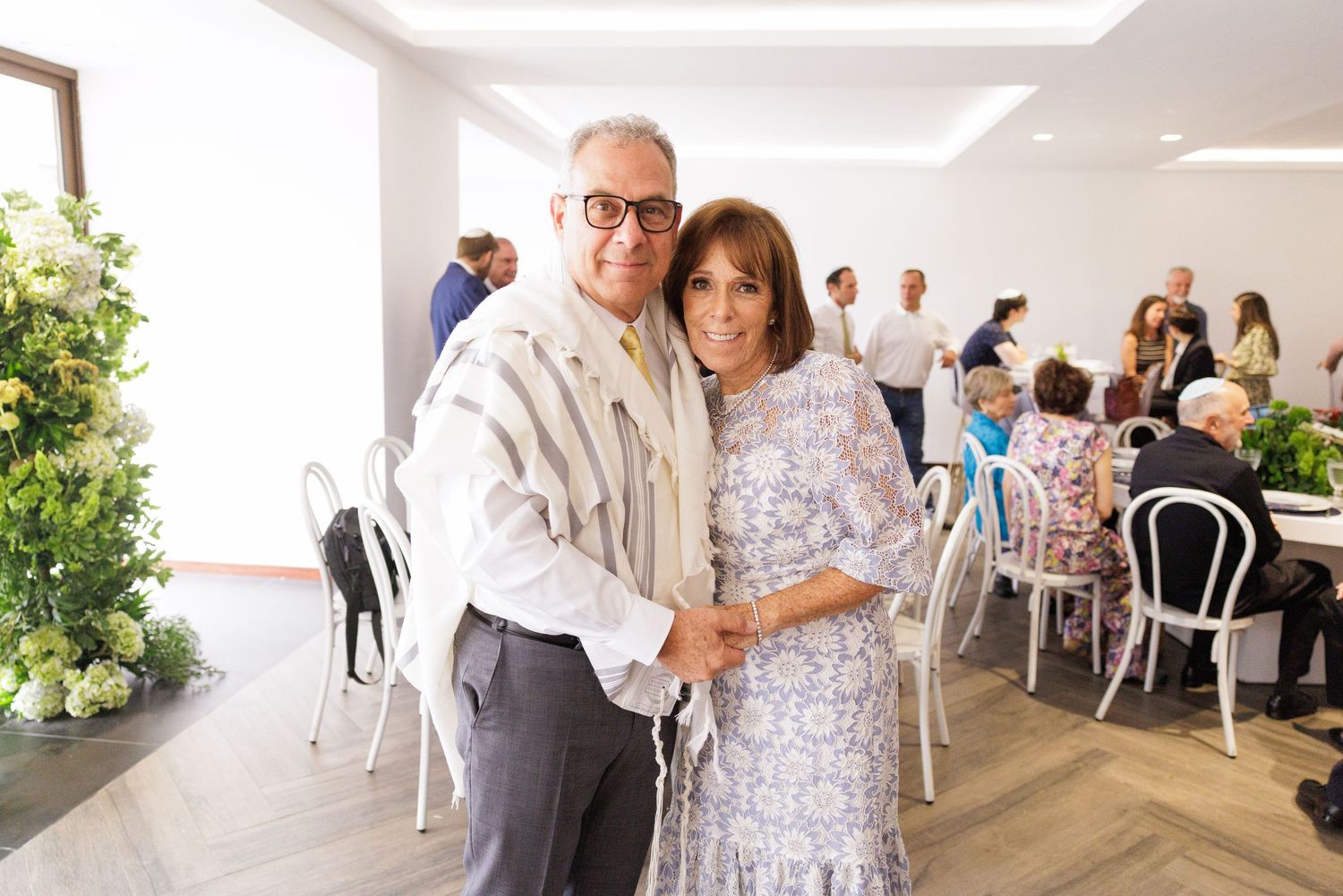 A couple embraces during a celebration in a modern venue with white chairs and tables.