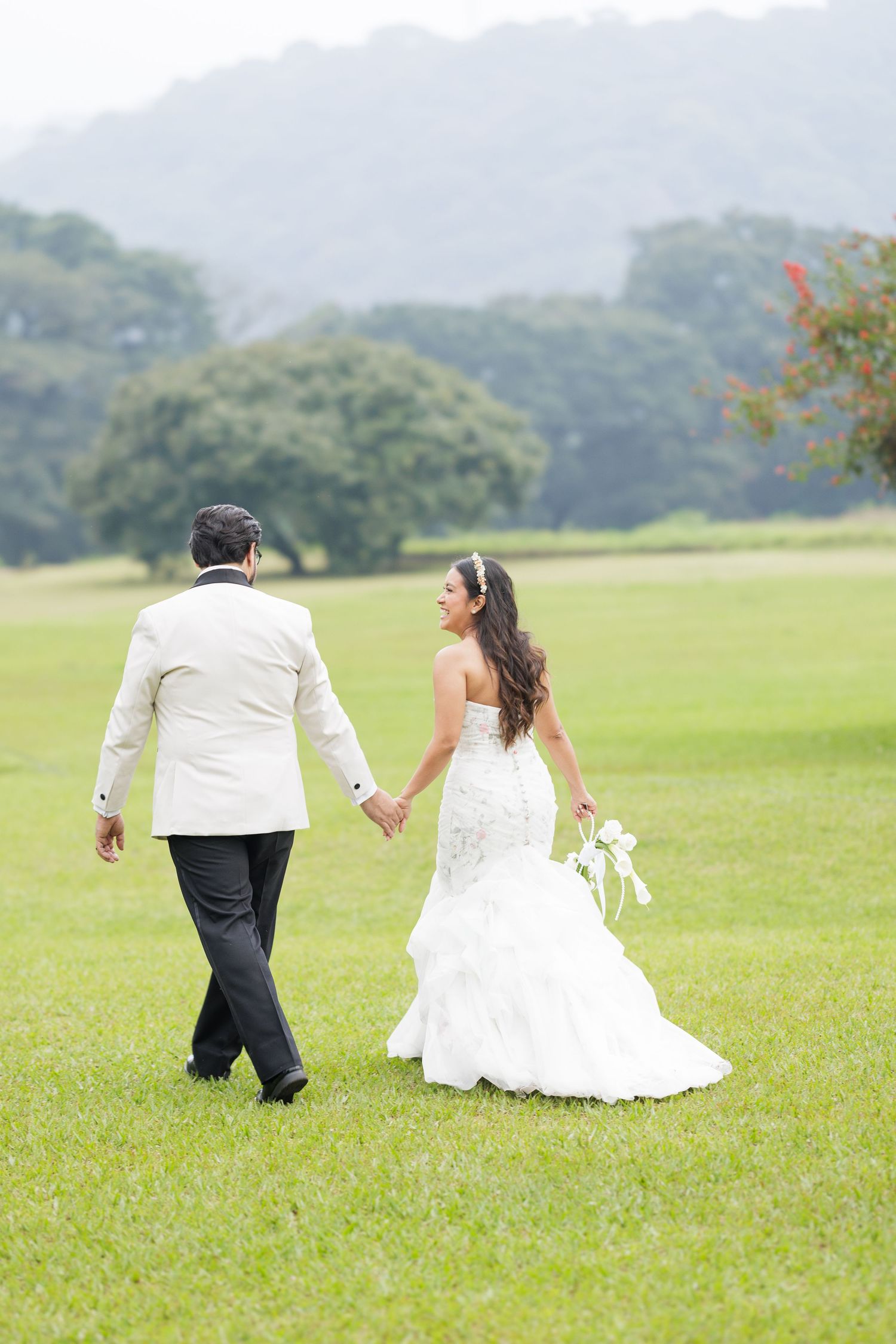 Bride and groom walk playfully across a vast green field with mountains in the background.