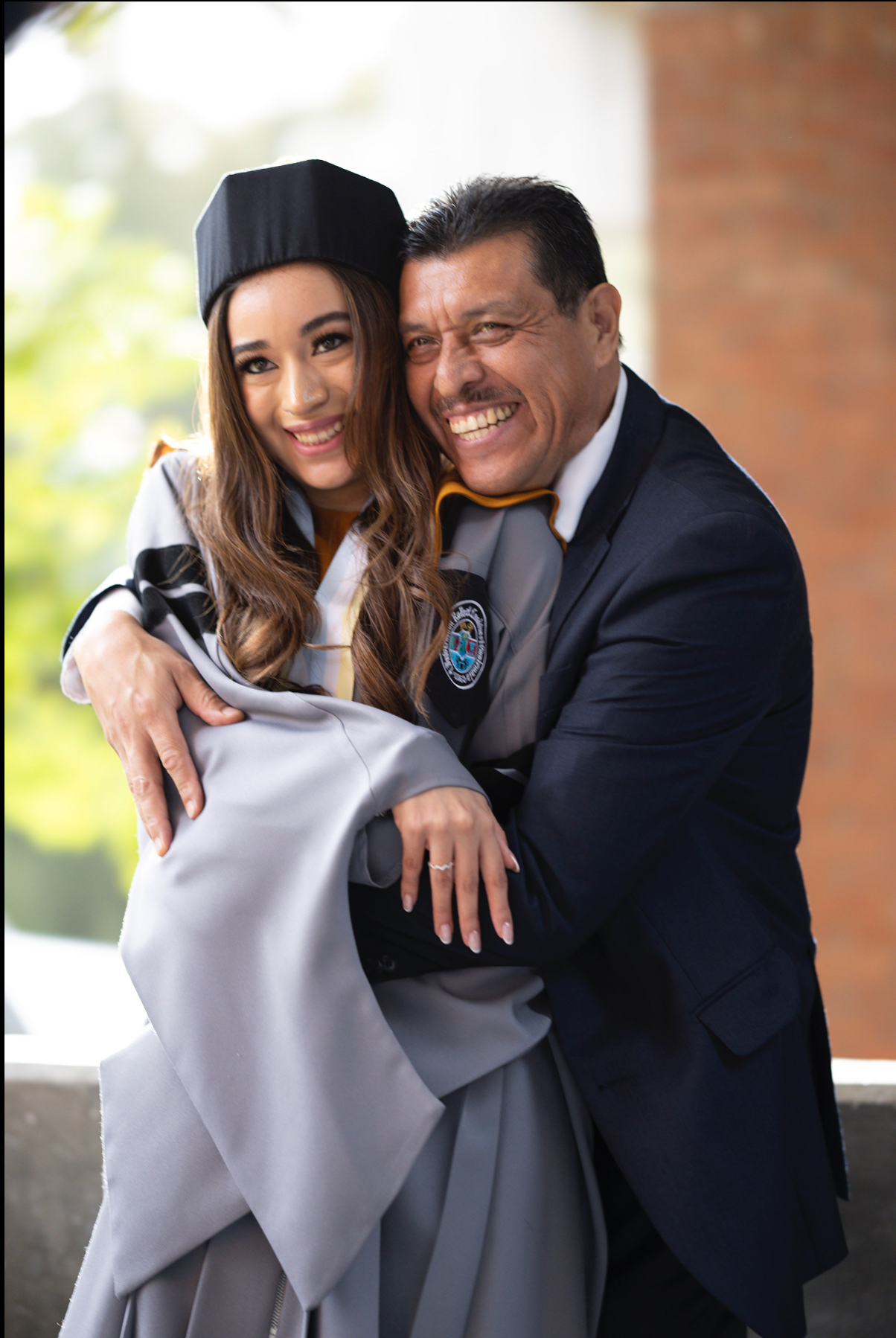 Happy graduates embrace while wearing formal attire and grey academic robes against brick wall.