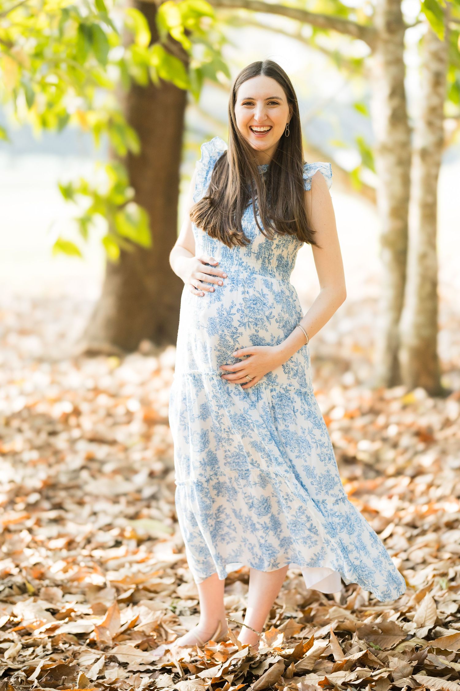 A blue and white floral dress twirls gracefully among fallen autumn leaves in a sunlit wooded setting.