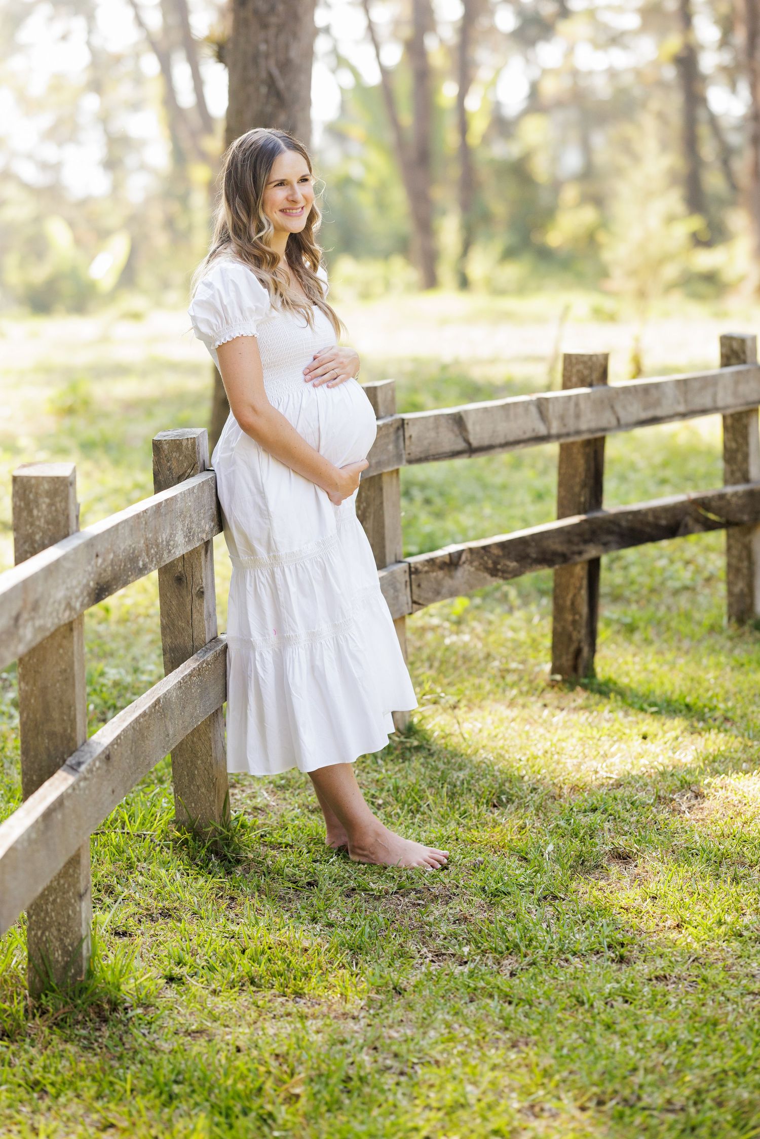 Woman in white dress poses along rustic wooden fence in sunlit forest.