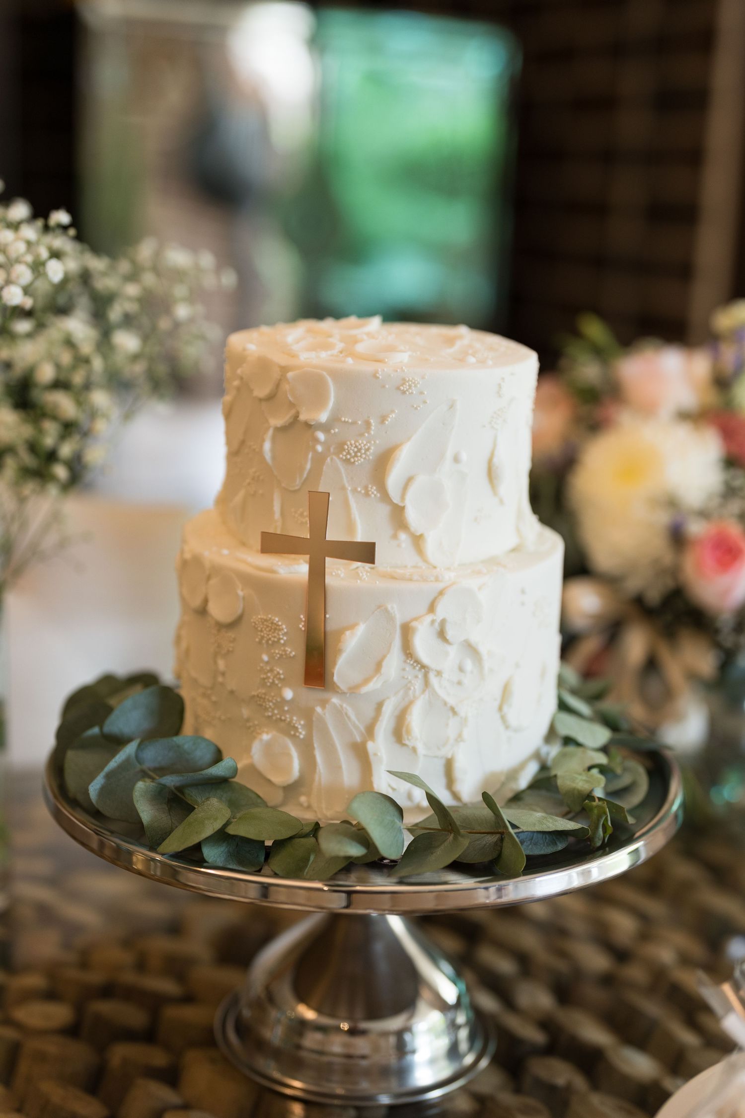 Elegant white wedding cake with delicate floral designs displayed on a silver cake stand with eucalyptus leaves.