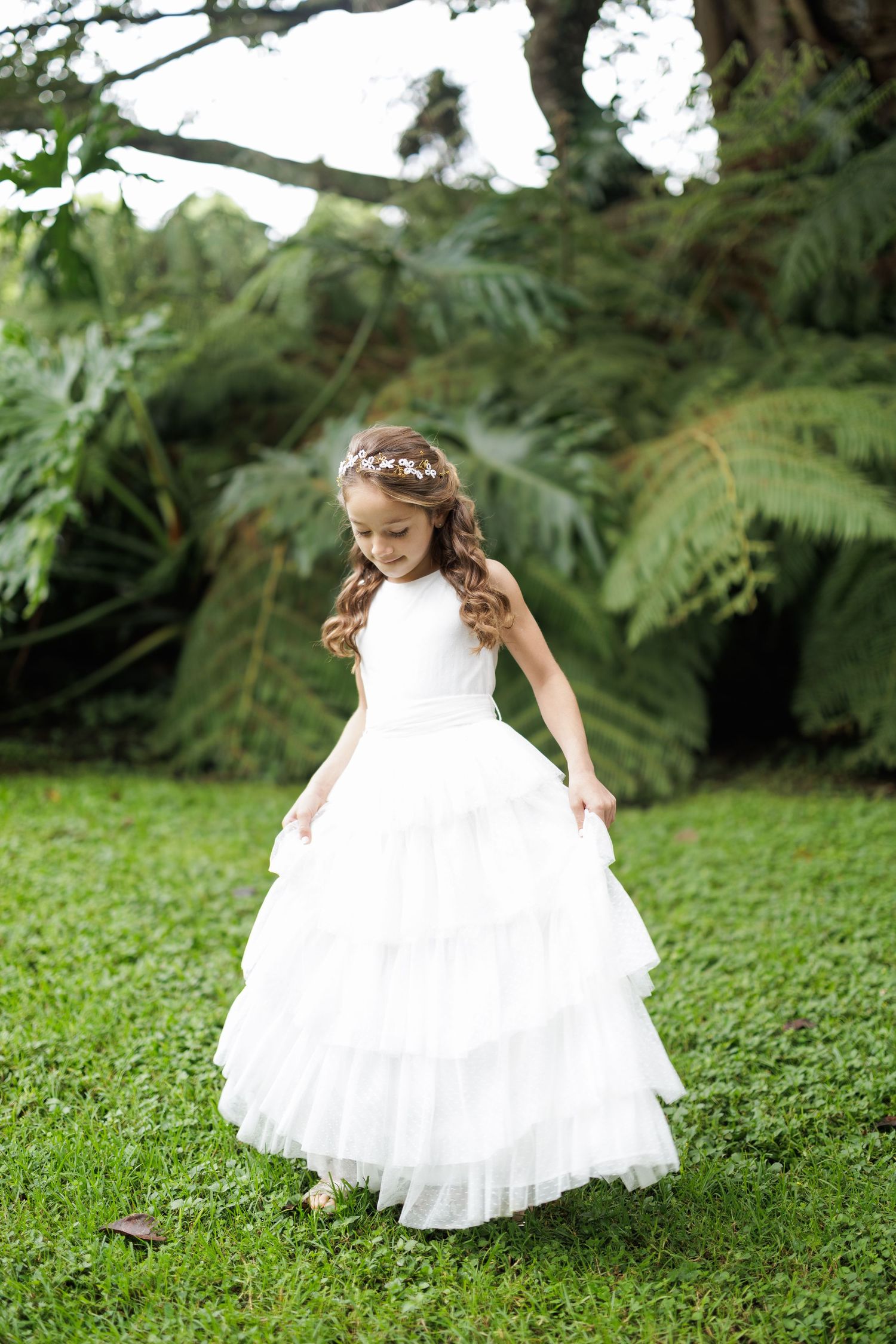 Young child in a flowing white dress twirls playfully on green grass surrounded by ferns.