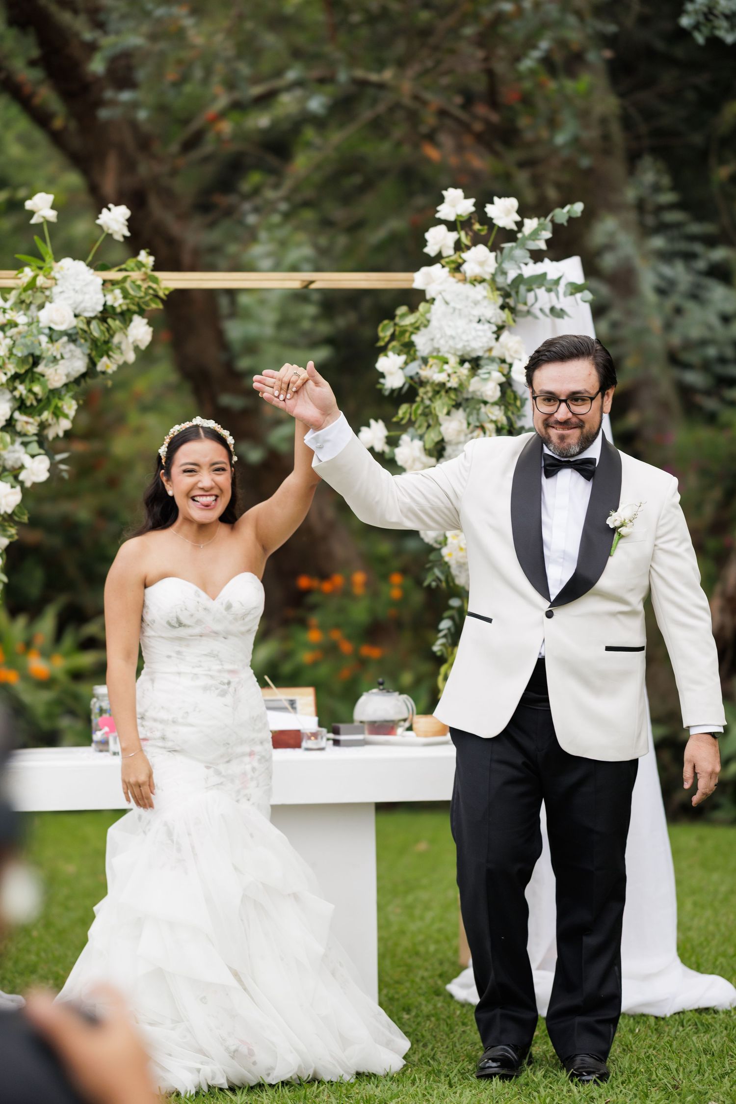Wedding couple shares playful moments on grass near white floral arrangements.