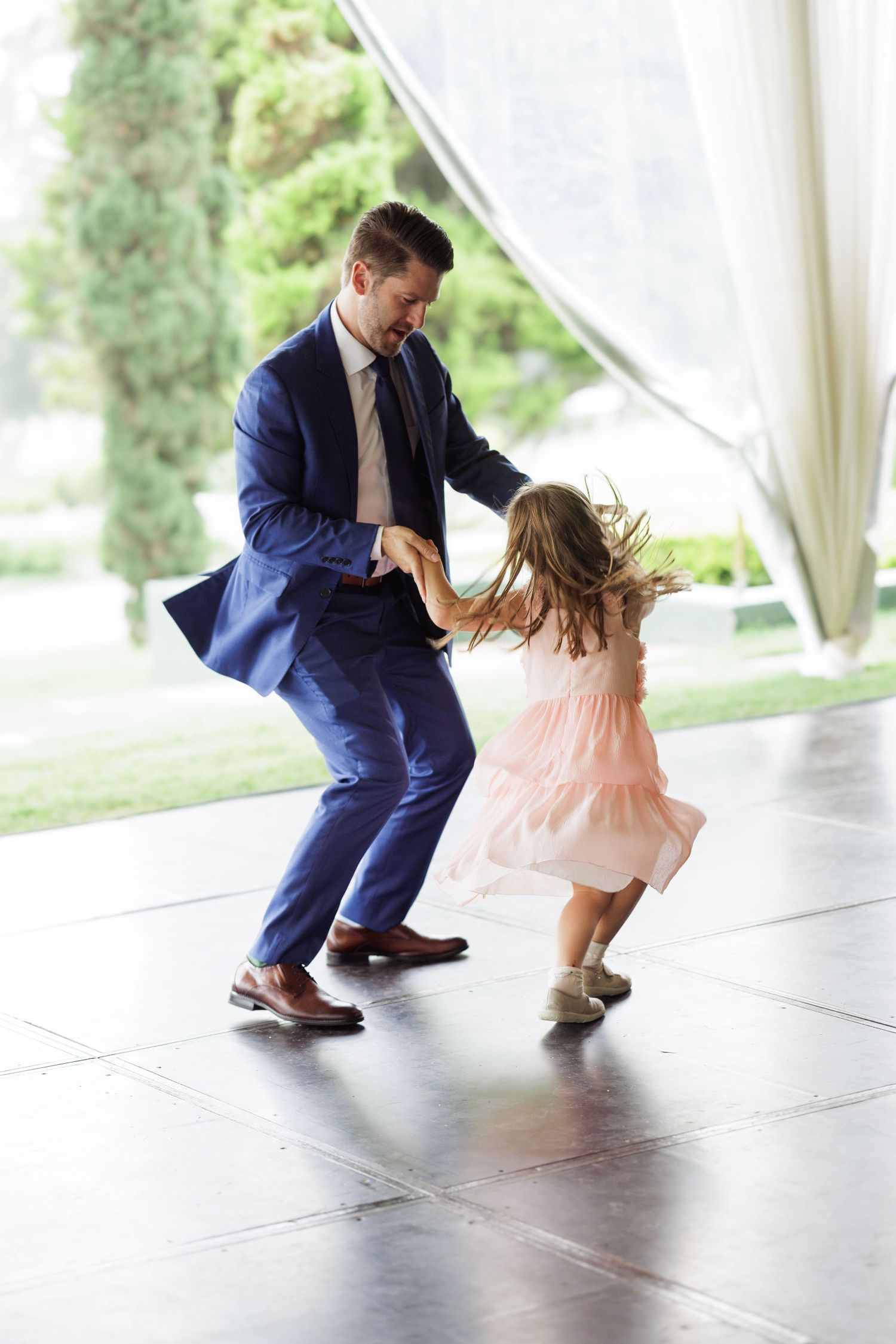 A sequence of dance moves between a person in a navy suit and small child in a pink dress on a covered patio.