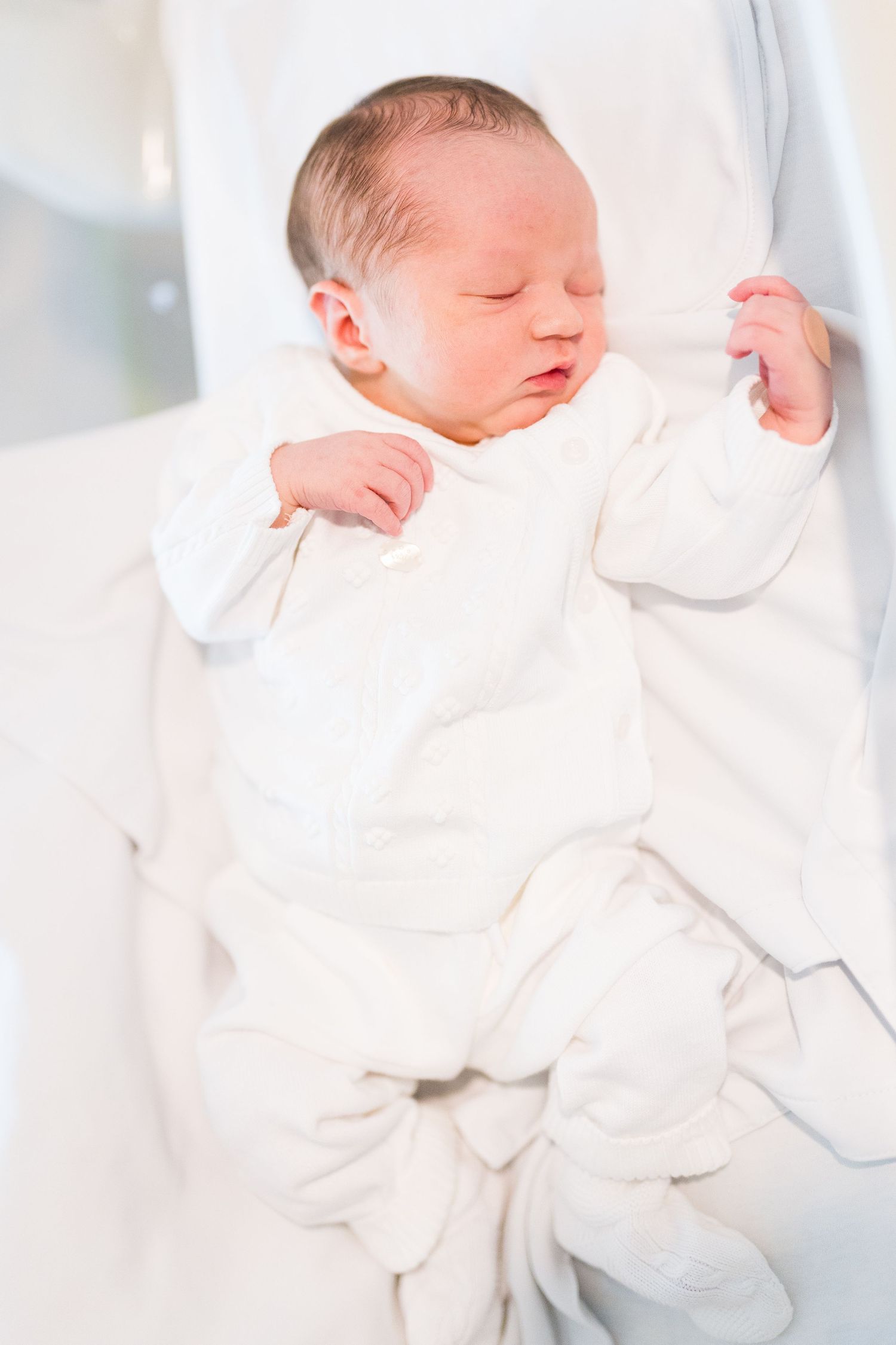 Newborn baby wearing white sleeps peacefully on soft white bedding.