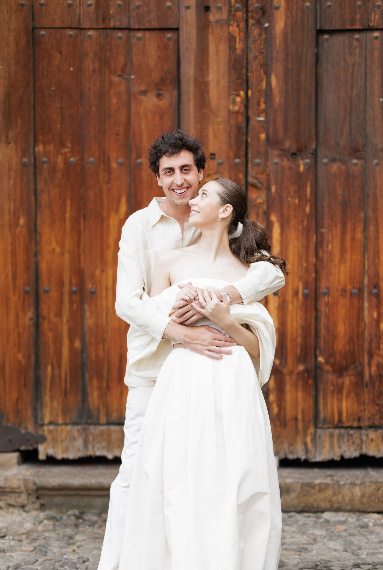 Romantic couple in white outfits shares intimate moment against wooden door.