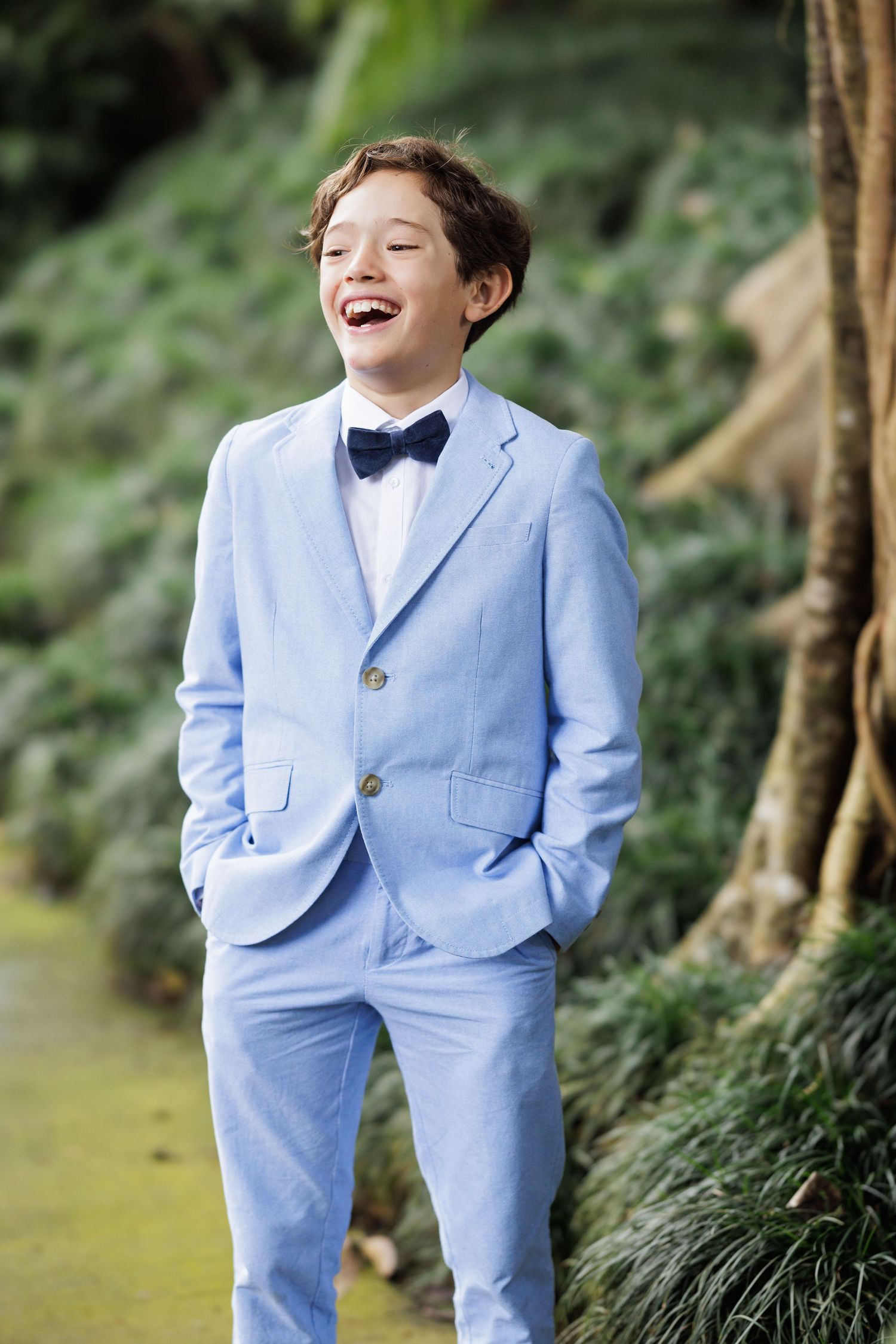 A child in a light blue suit poses playfully in a garden setting.