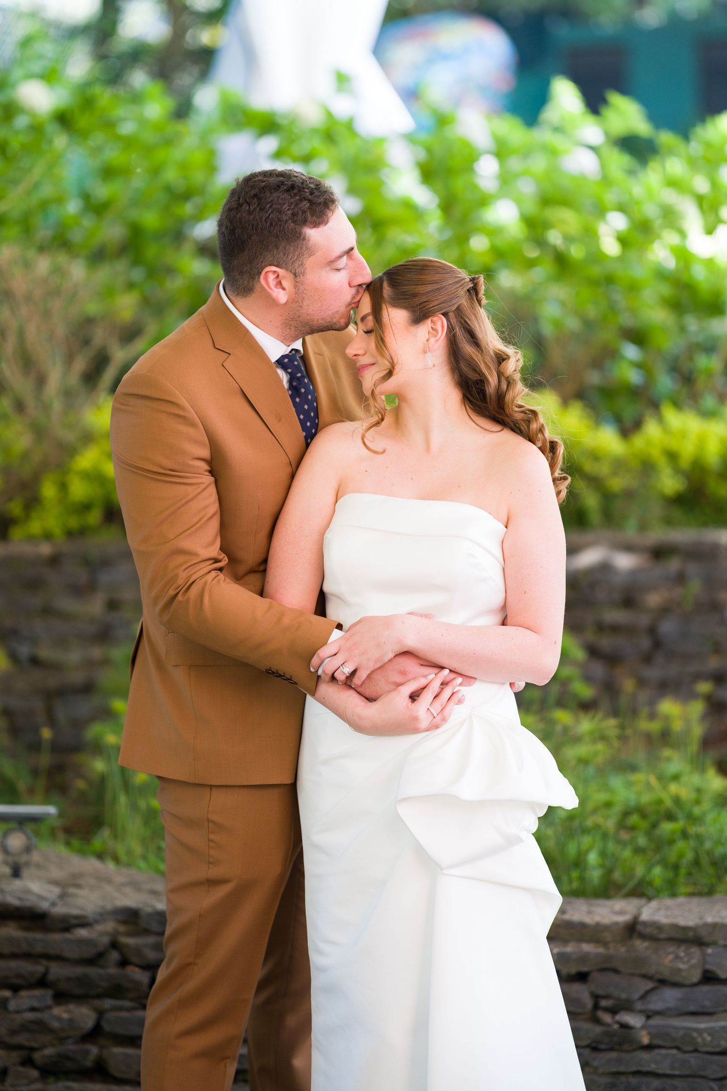 Romantic wedding couple embraces by stone wall in garden setting.