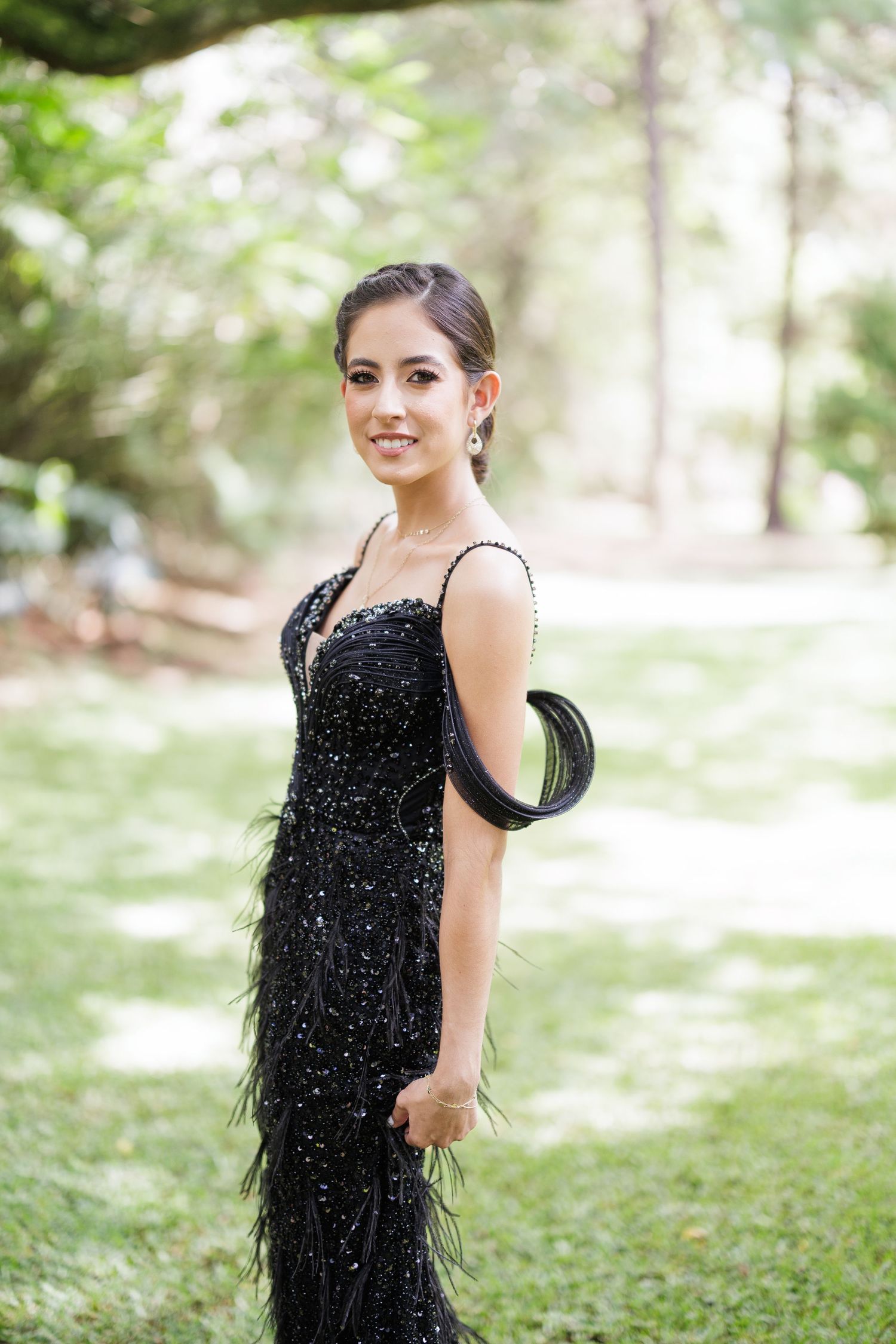 A person in a sparkling black formal dress poses elegantly in a sunlit outdoor setting.