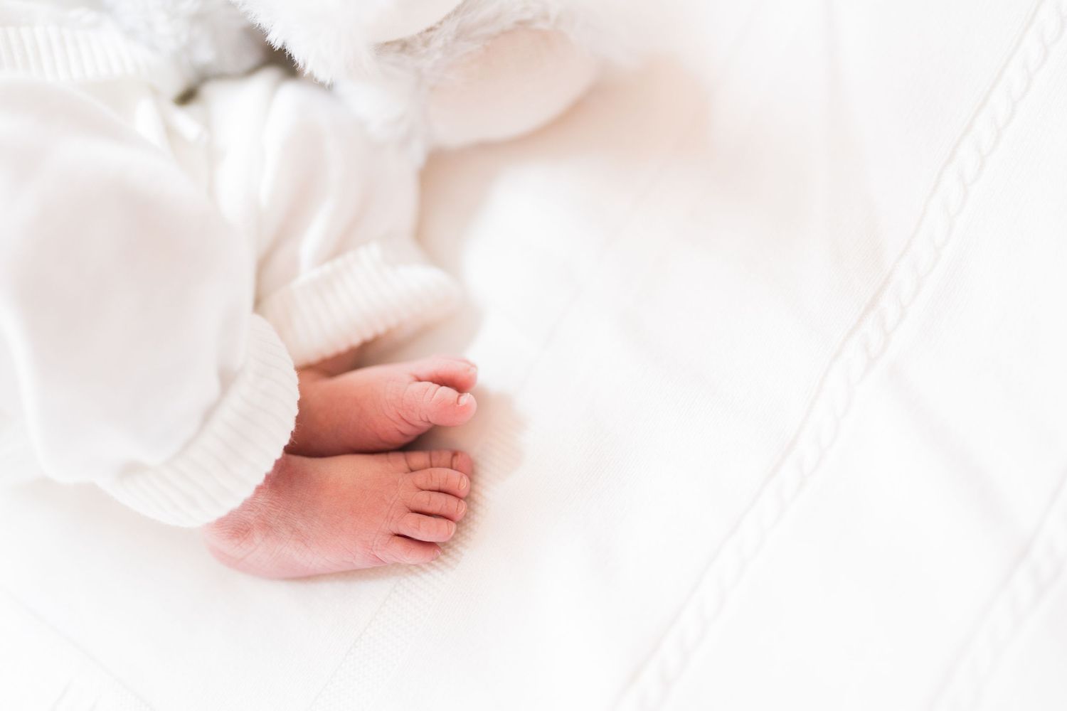 Newborn baby in white clothing sleeping next to a plush teddy bear on white bedding.