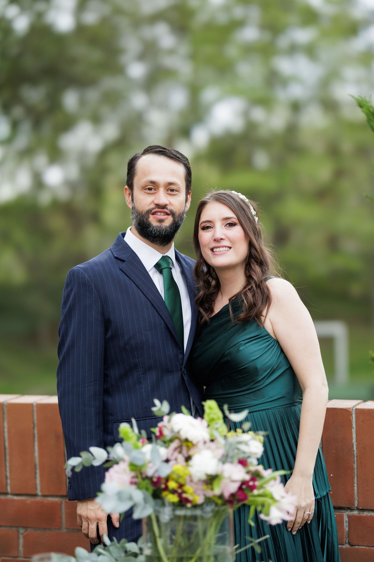 A couple poses together outdoors against a brick wall with floral arrangements.