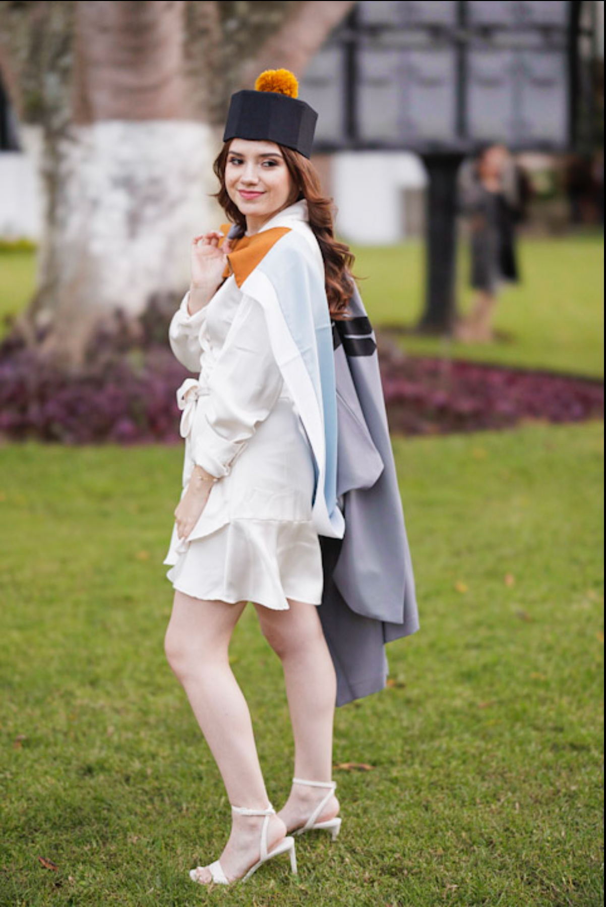Graduate in white dress and grey academic gown poses on grass.