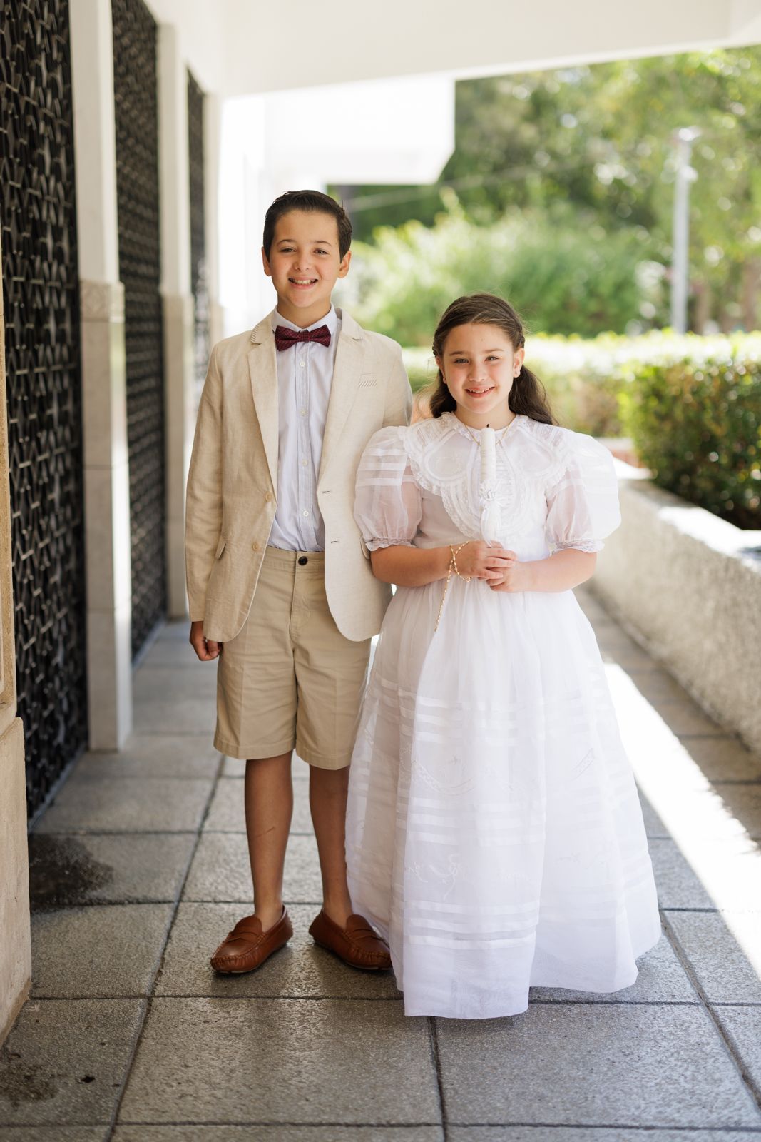 Young boy in tan suit and girl in white dress pose formally on outdoor walkway.