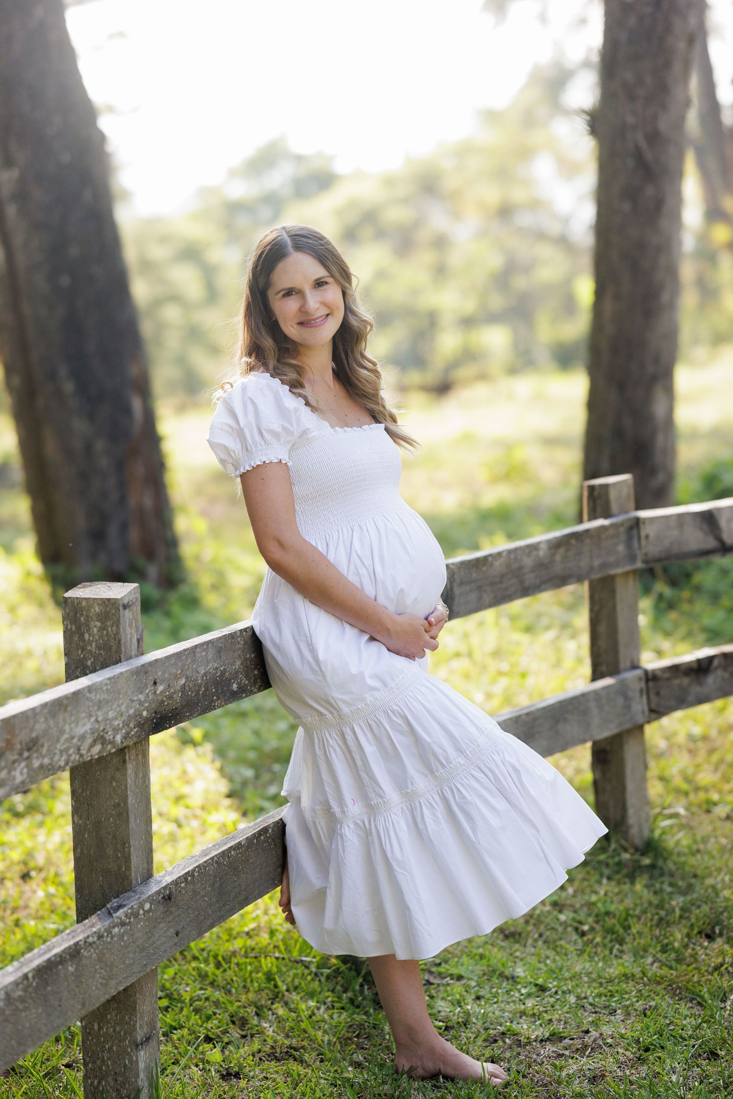 A white dress flows gracefully against a rustic wooden fence in a sunny outdoor setting.